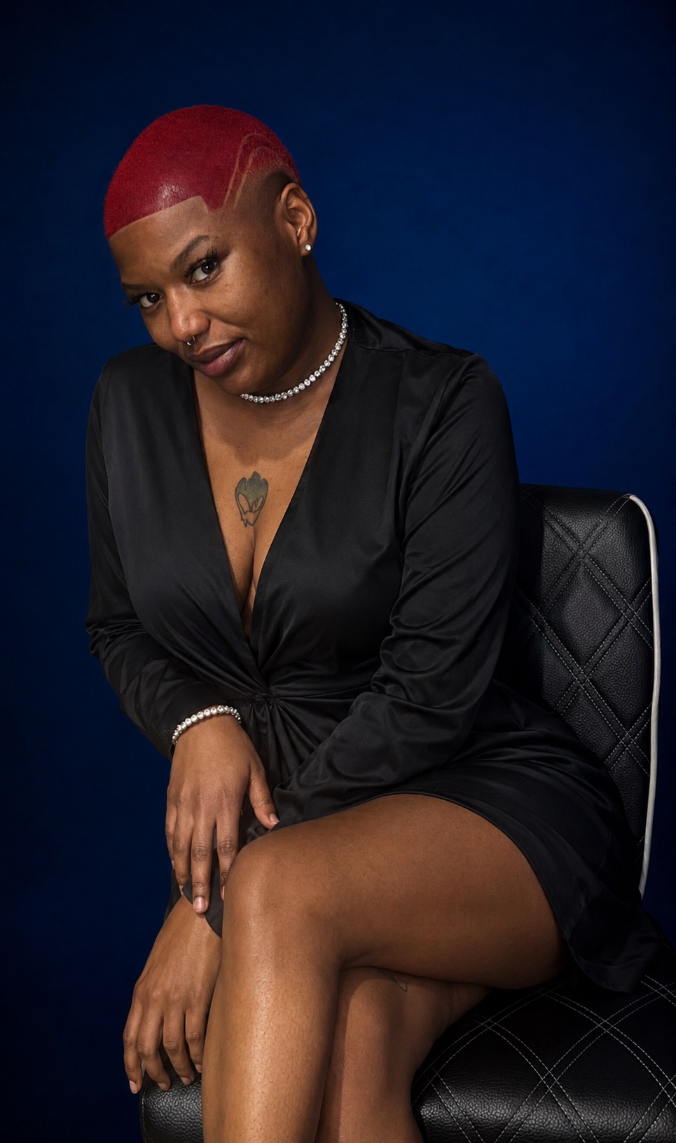 Woman with red hair sitting on a chair, wearing a black dress, against a dark blue background.