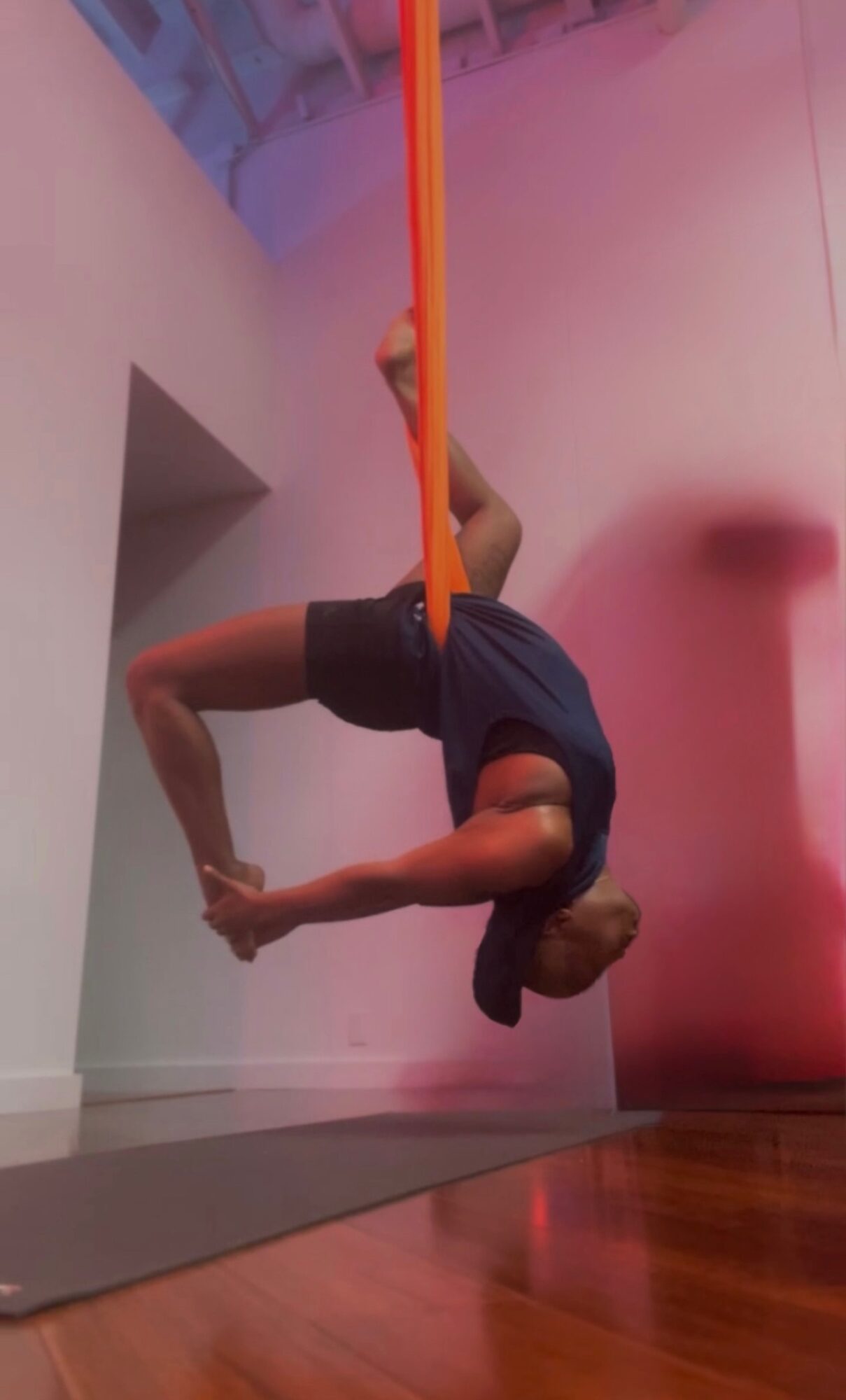 Person hanging upside down from a yellow and orange aerial silk, in an indoor space with pink and white walls.