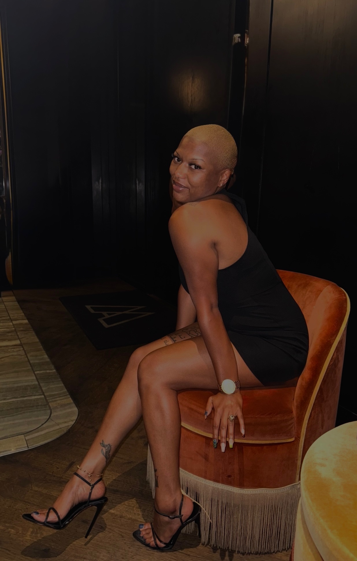 Woman with a shaved head sitting on a round orange chair in a black dress and high heels, looking at the camera.