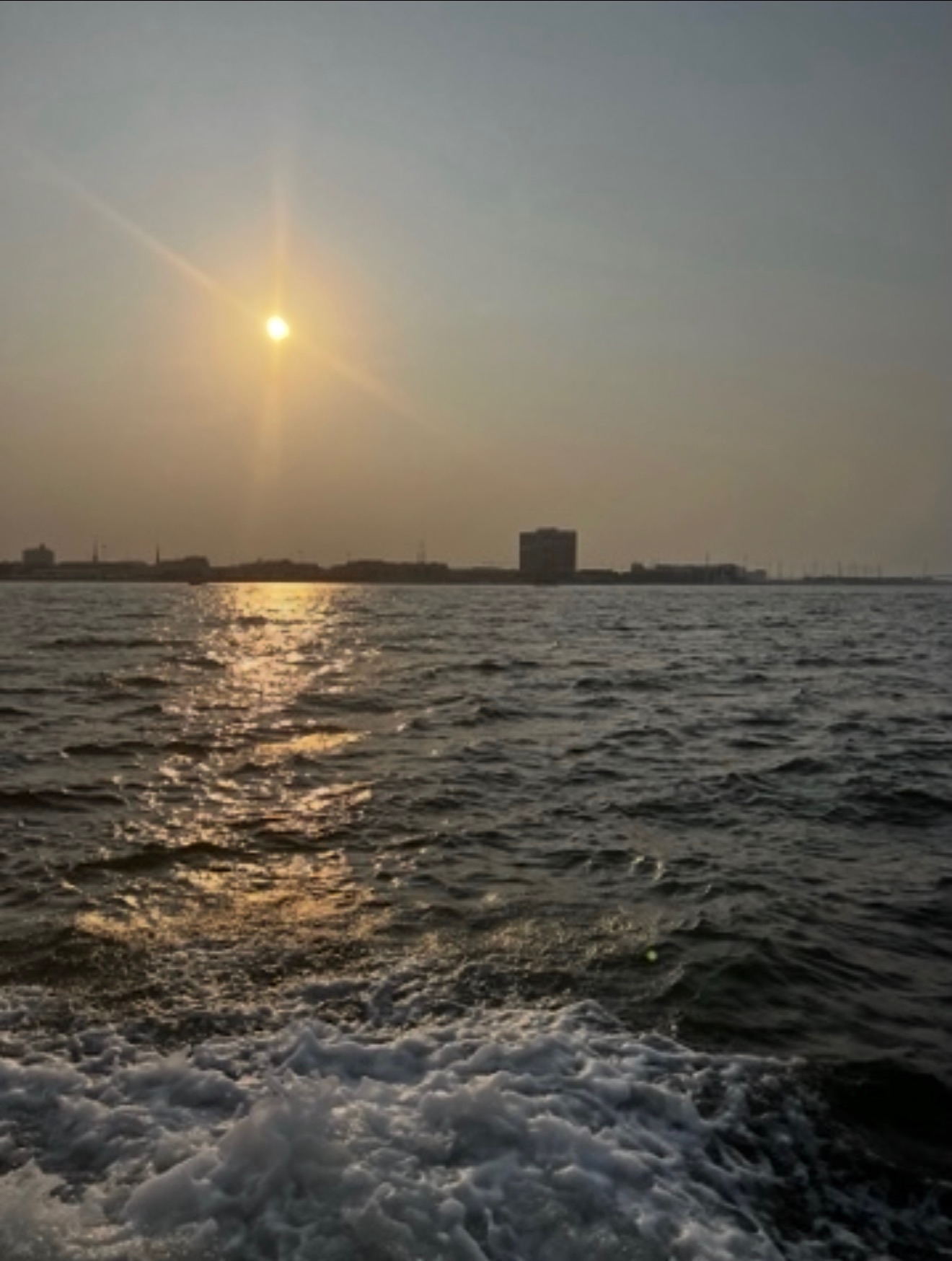 Sunset over water with buildings in the distance, water reflecting sunlight, sky clear with a few clouds, calm water surface.