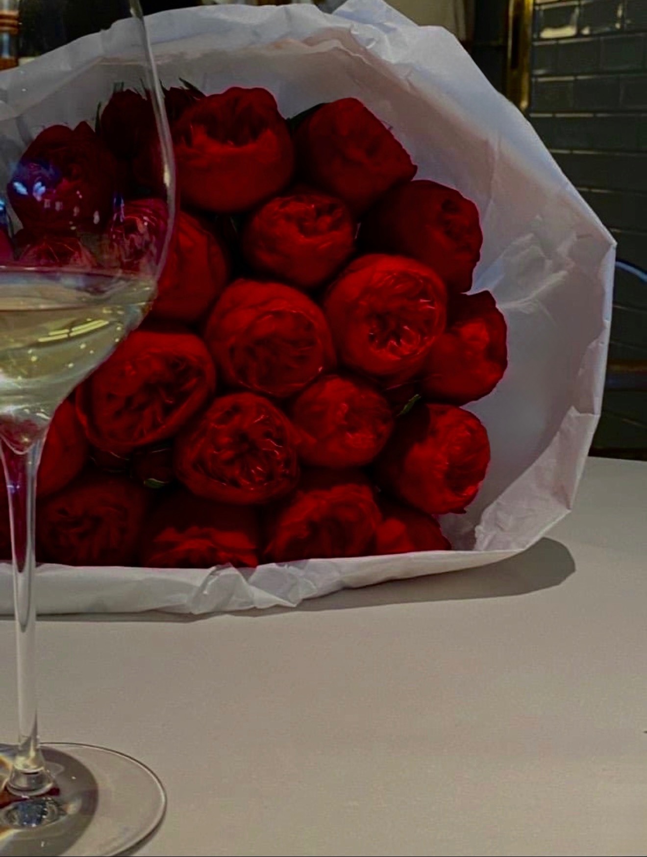 Bouquet of red roses wrapped in white paper with a glass of white wine nearby on a table.