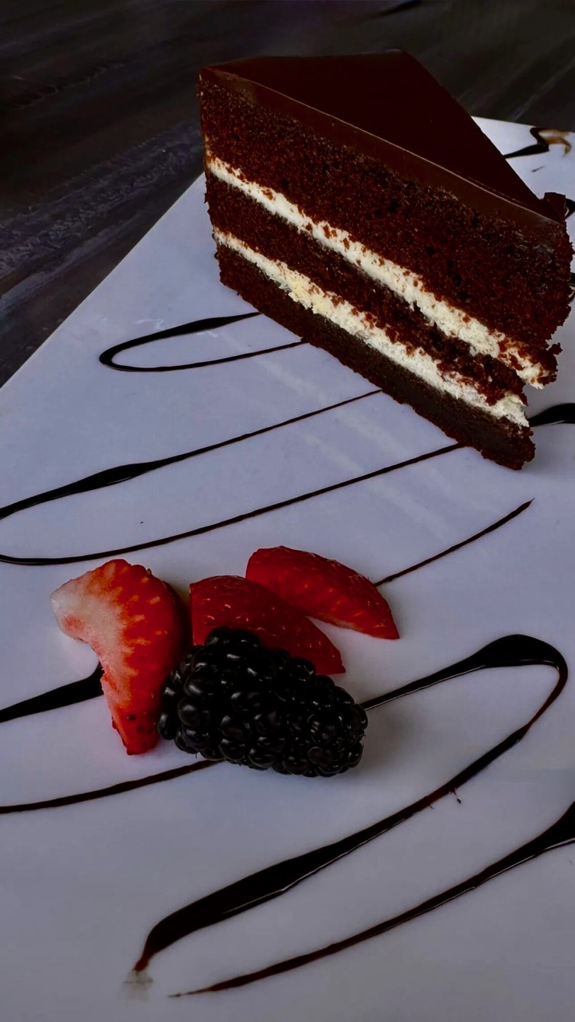 Slice of red velvet cake with cream filling on a white plate, garnished with strawberries and blackberries, with chocolate drizzle.