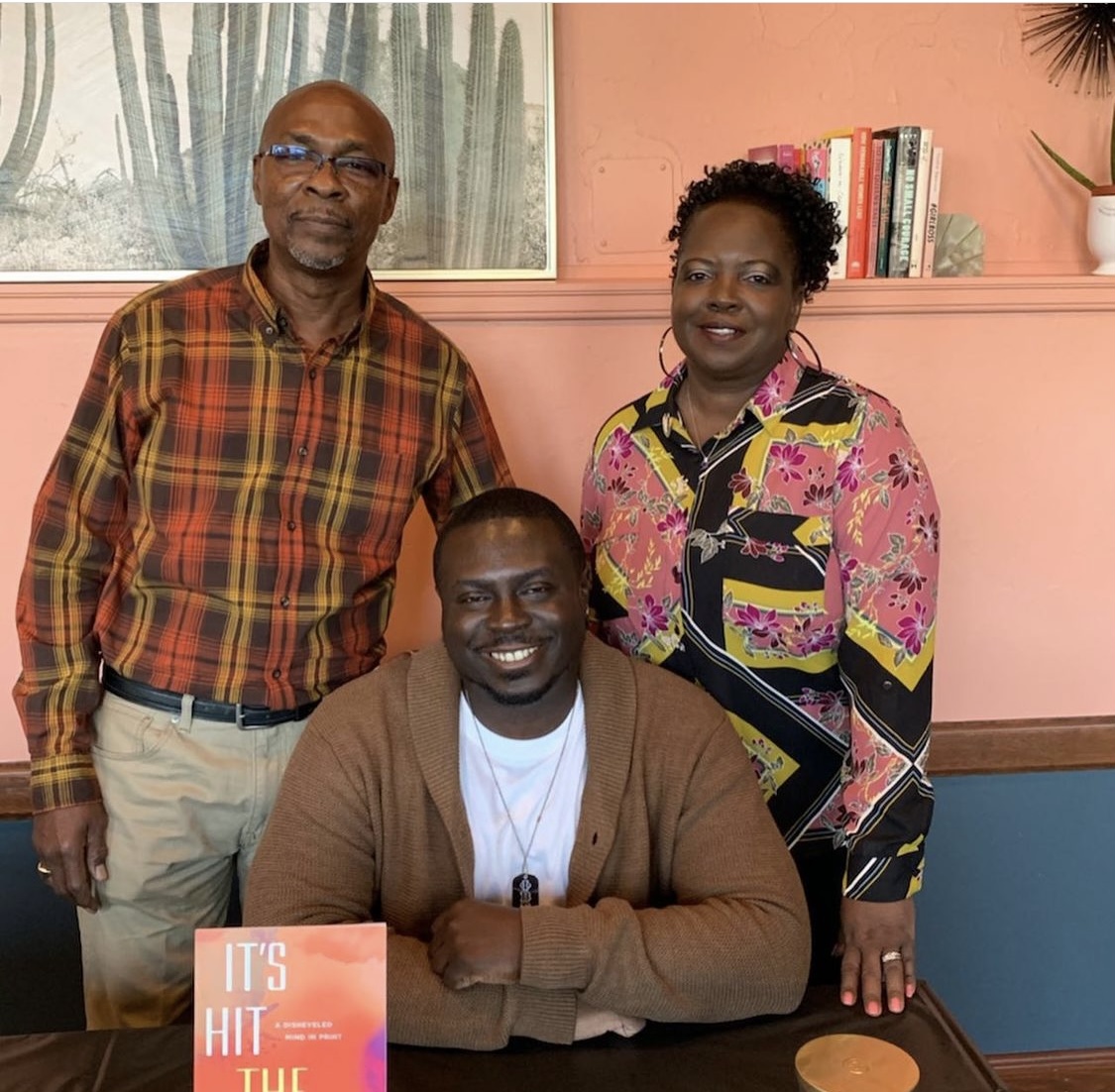 Three people, two standing and one sitting, in a room with pink and blue walls, books, and a plant.