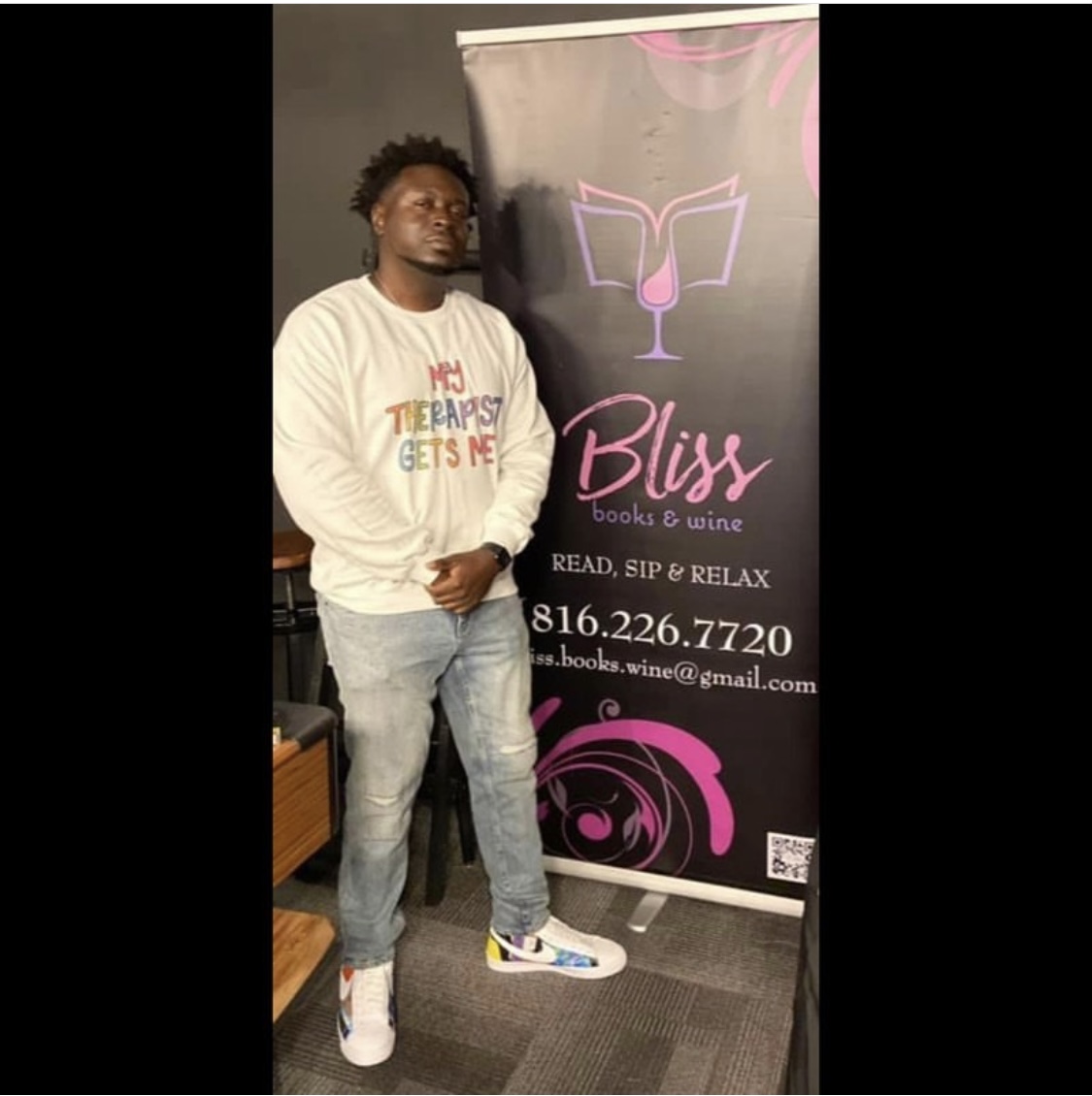 Young man standing next to a banner for Bliss books & wine, holding a phone, wearing a white sweatshirt and jeans.