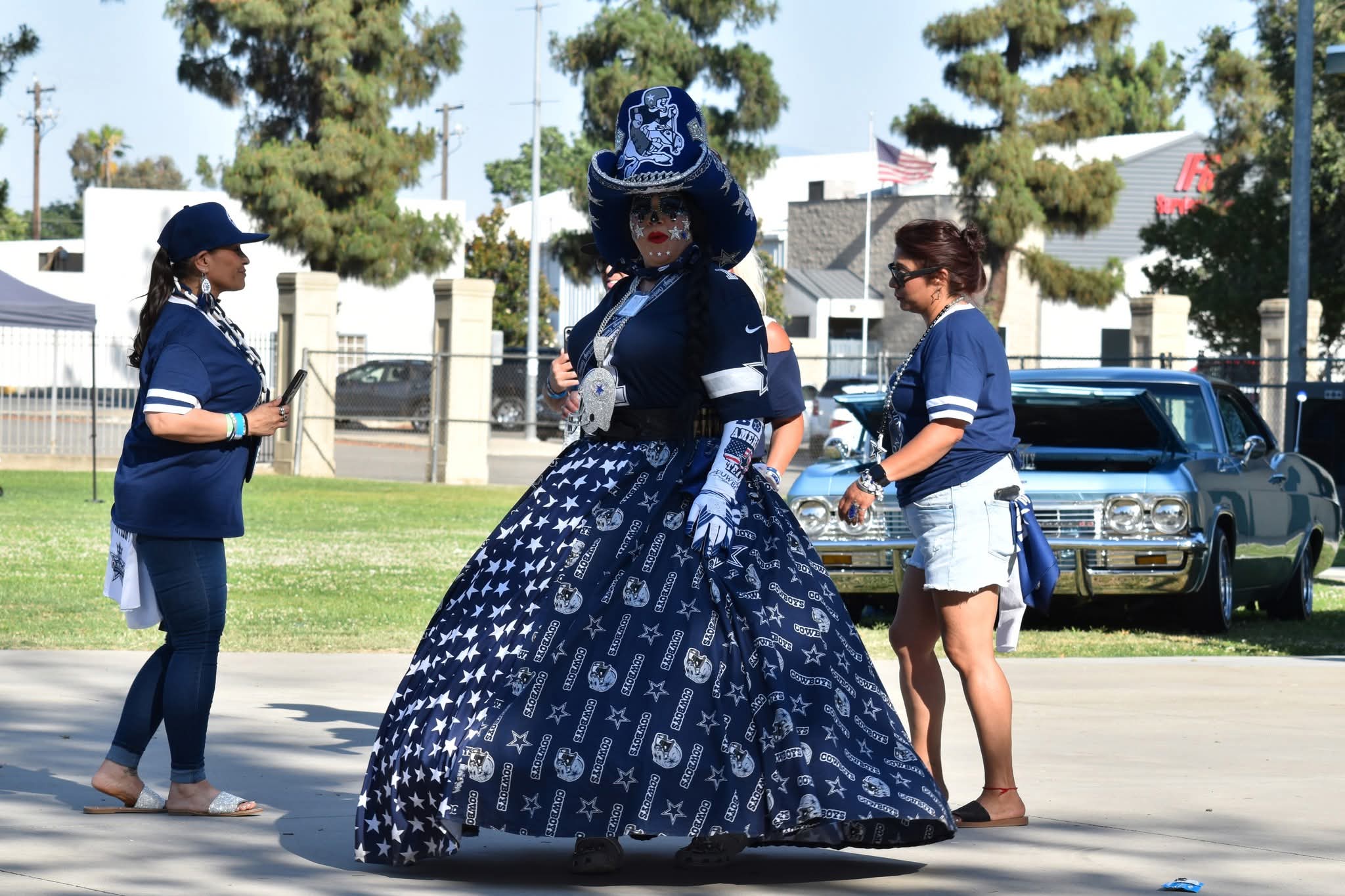 Person in elaborate blue dress and hat, surrounded by three women in casual clothing, outdoors on pavement with cars and trees in background.