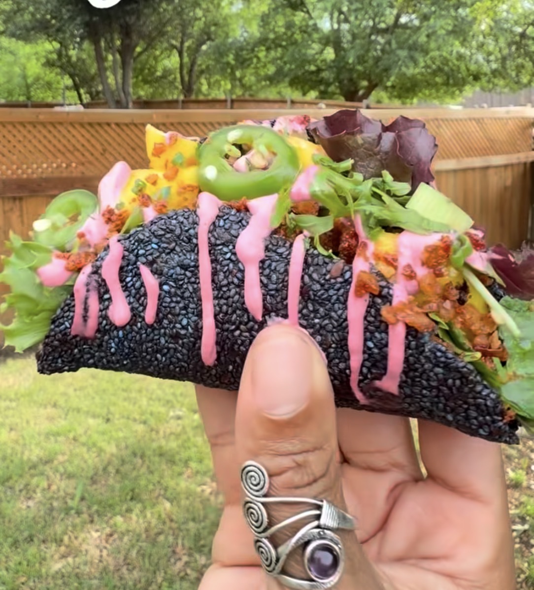 Hand holding a black taco shell with colorful toppings, including lettuce, cheese, and pink sauce, outdoors with trees and grass.
