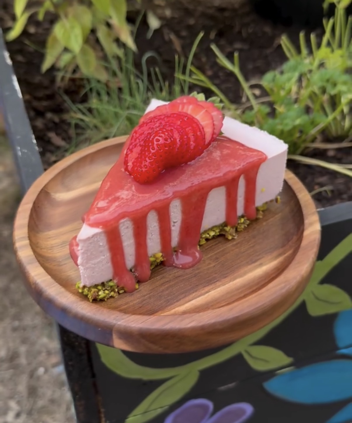 Slice of strawberry cheesecake with strawberries on top, drizzled with pink sauce, on a wooden plate, outdoors.