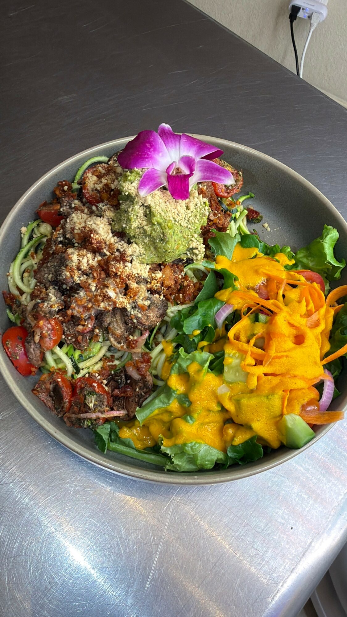 Salad with greens, cherry tomatoes, shredded cheese, purple orchid flower, and yellow shredded topping in a bowl on a dark surface.