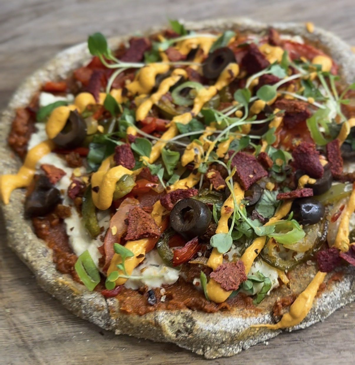 Vegetarian pizza topped with black olives, greens, yellow sauce, and various vegetables on a rustic wooden surface.