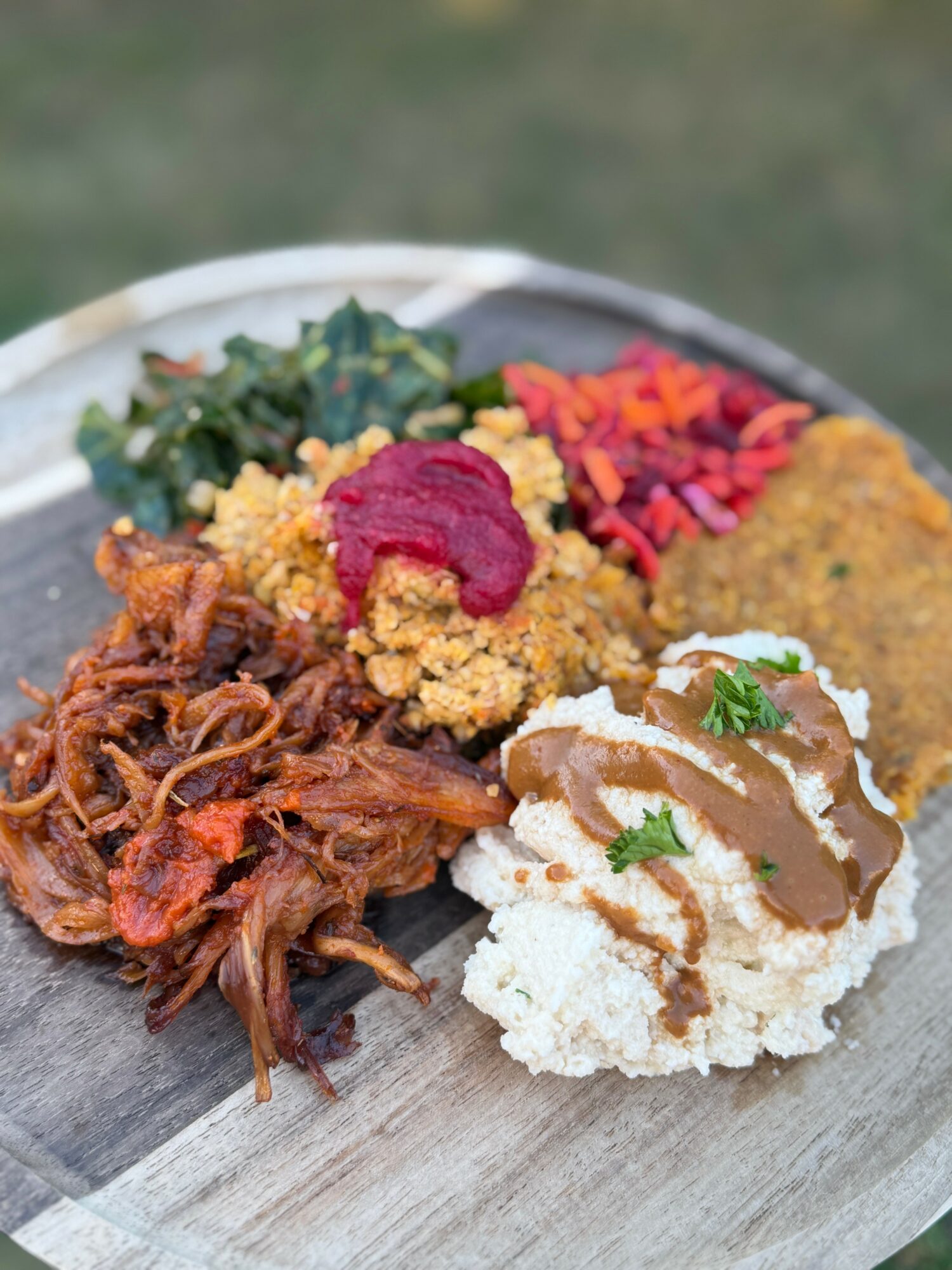 Plate with shredded meat, mashed potatoes with gravy, vegetables, and bread with toppings, on a wooden surface.