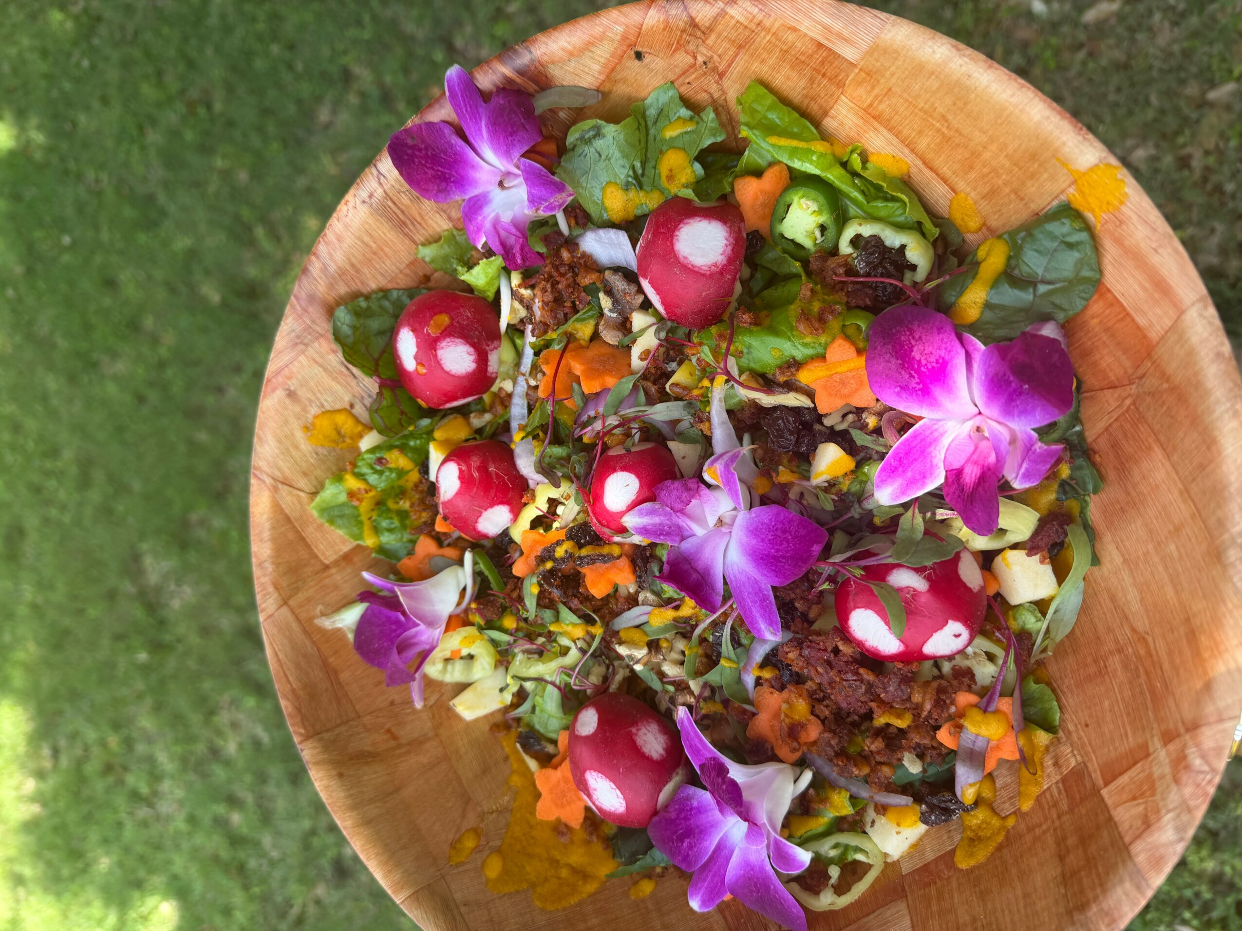 Colorful salad with purple flowers, mushrooms, and greens in a wooden bowl on grass.