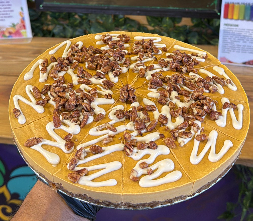 A round cake topped with caramel, chopped nuts, and white icing writing on top, on a wooden surface.