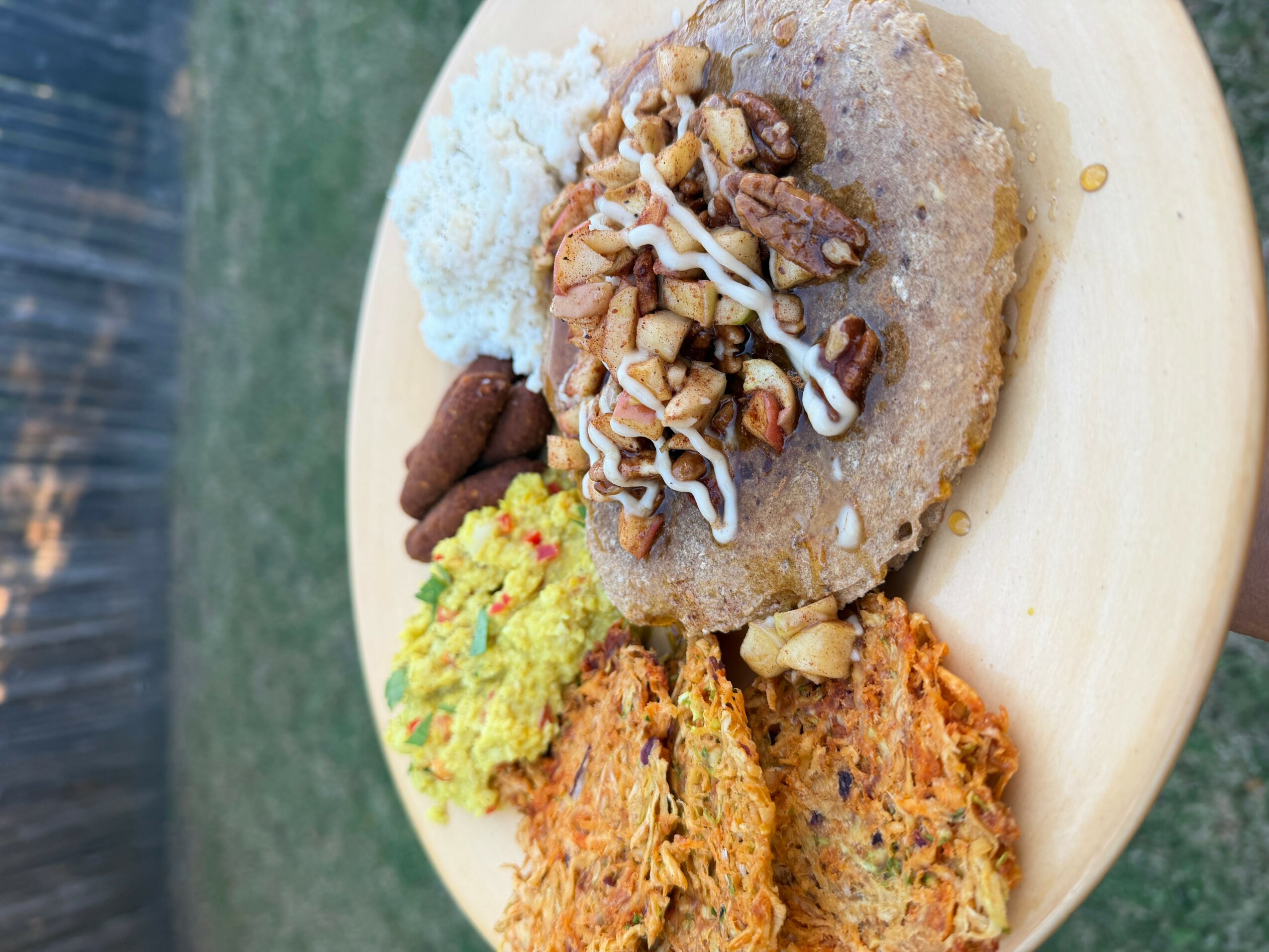 Plate with a large, round, fried snack topped with nuts, shredded coconut, and chocolate, alongside green and orange side dishes.