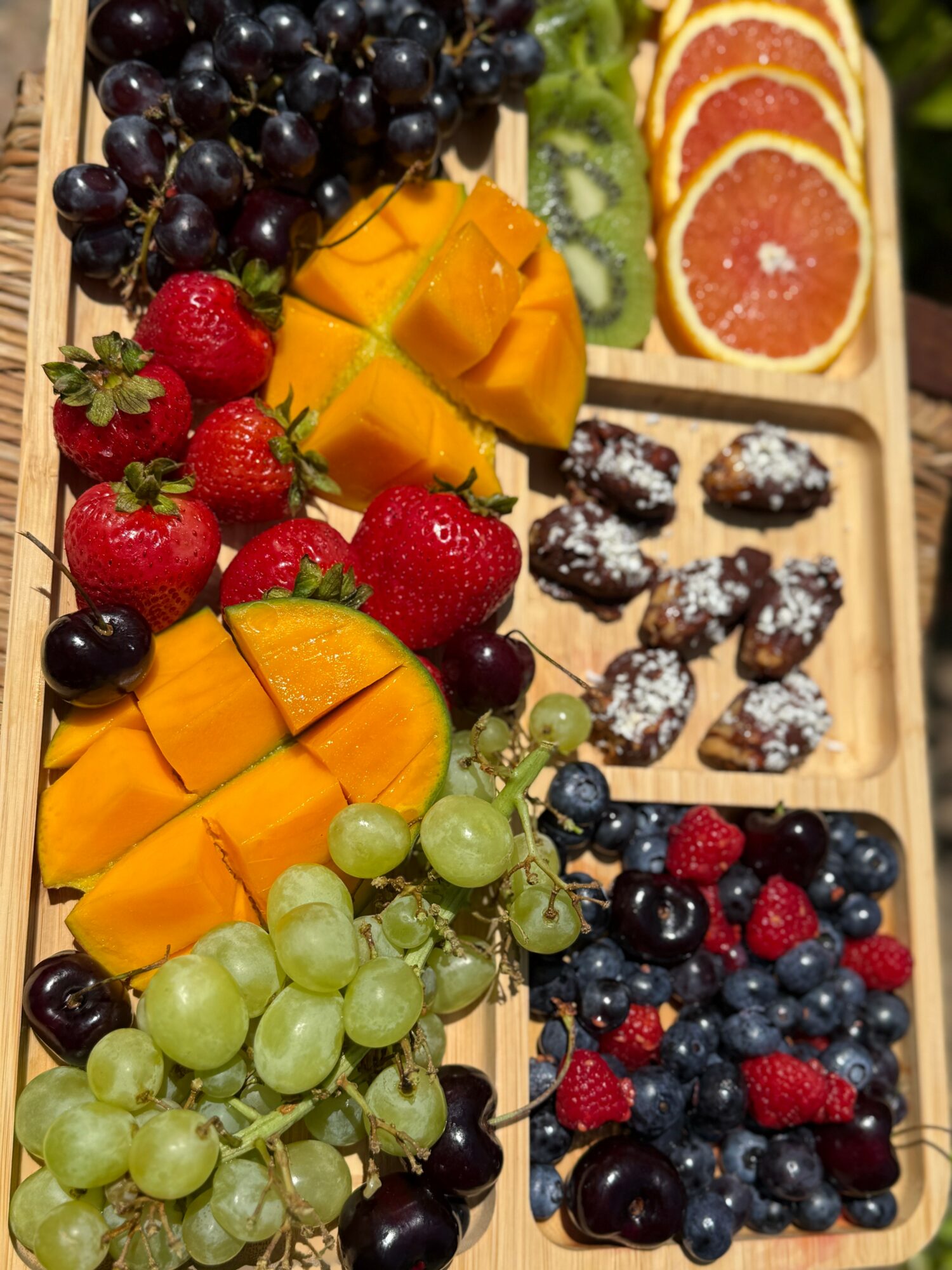 Assorted fresh fruits including grapes, strawberries, mango, orange slices, and berries in a wooden tray.