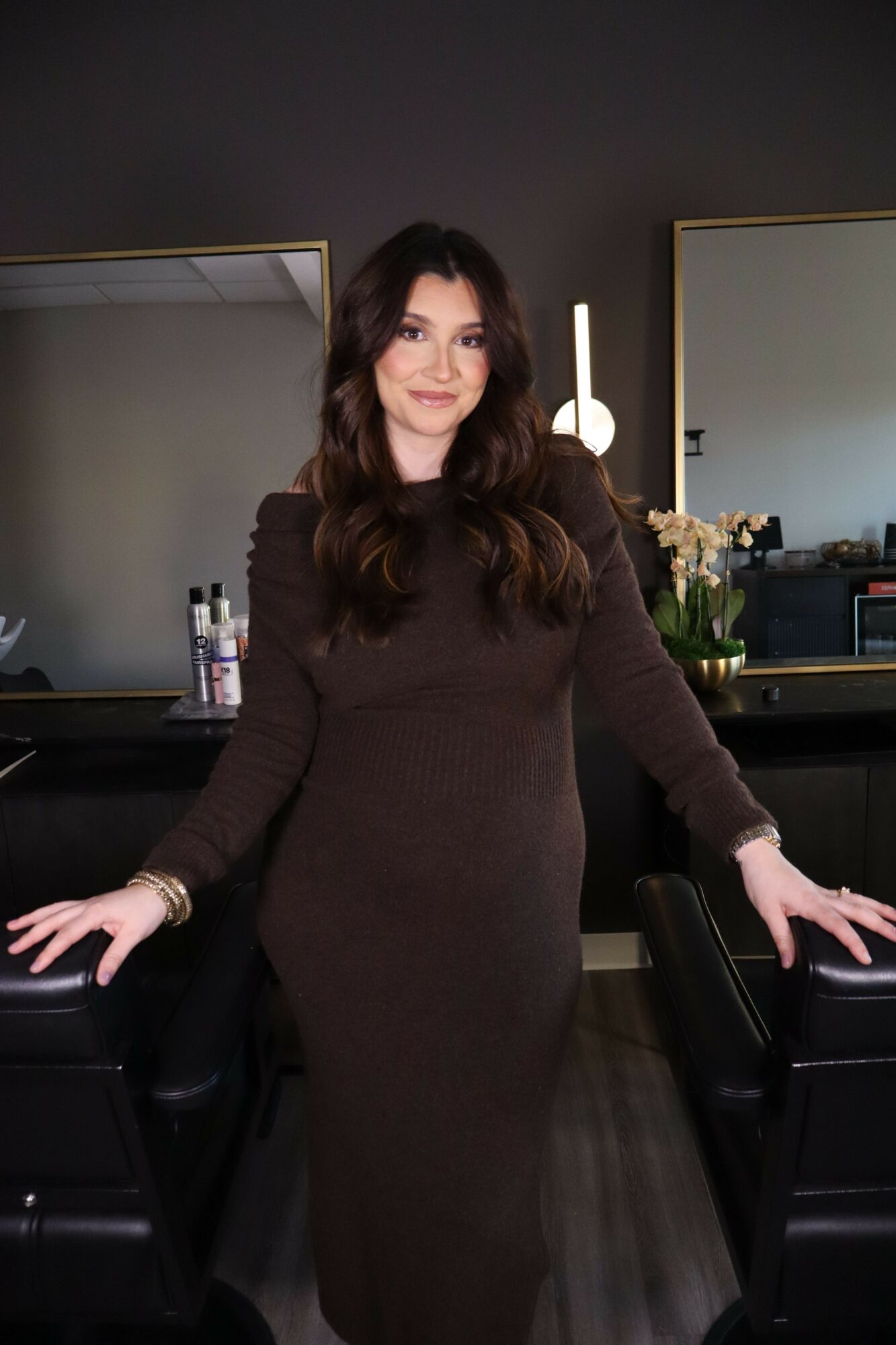 Woman with long dark hair in a brown dress standing in a room with dark walls, a mirror, and a desk, smiling and waving.