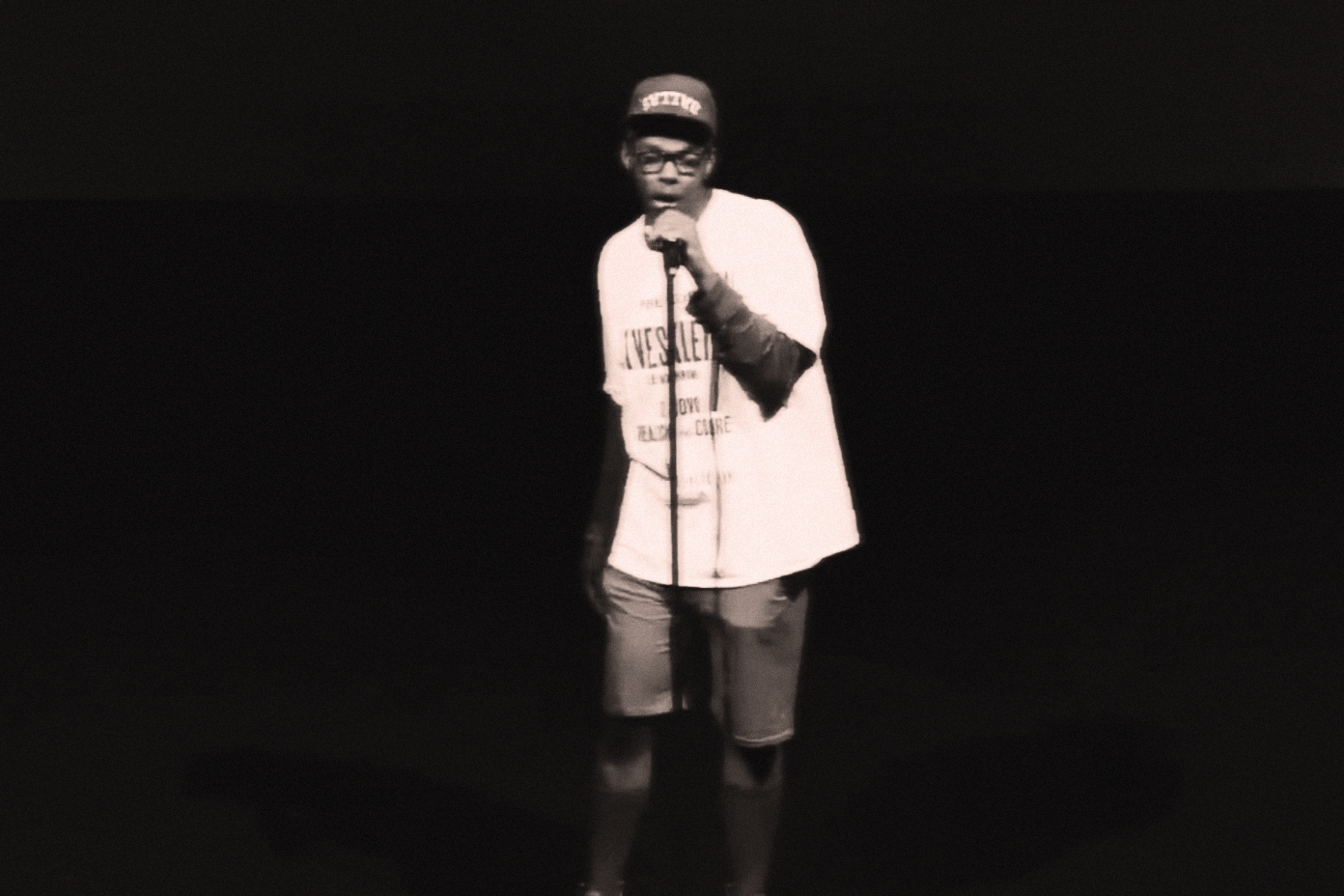 Person standing on stage holding a microphone, wearing a cap, t-shirt, and shorts, with a dark background.