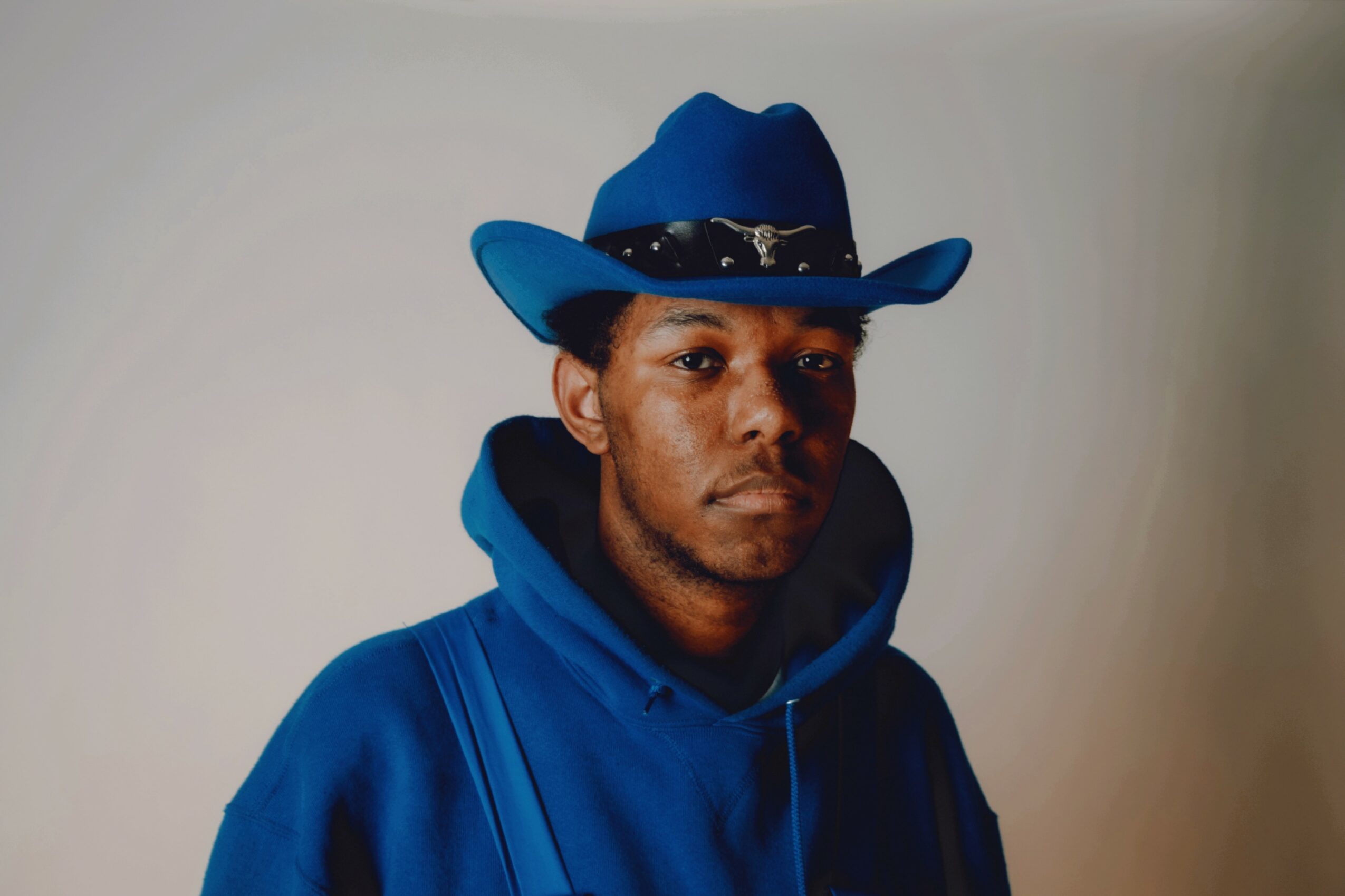 Young man wearing a blue cowboy hat and hoodie, looking serious, against a plain background.