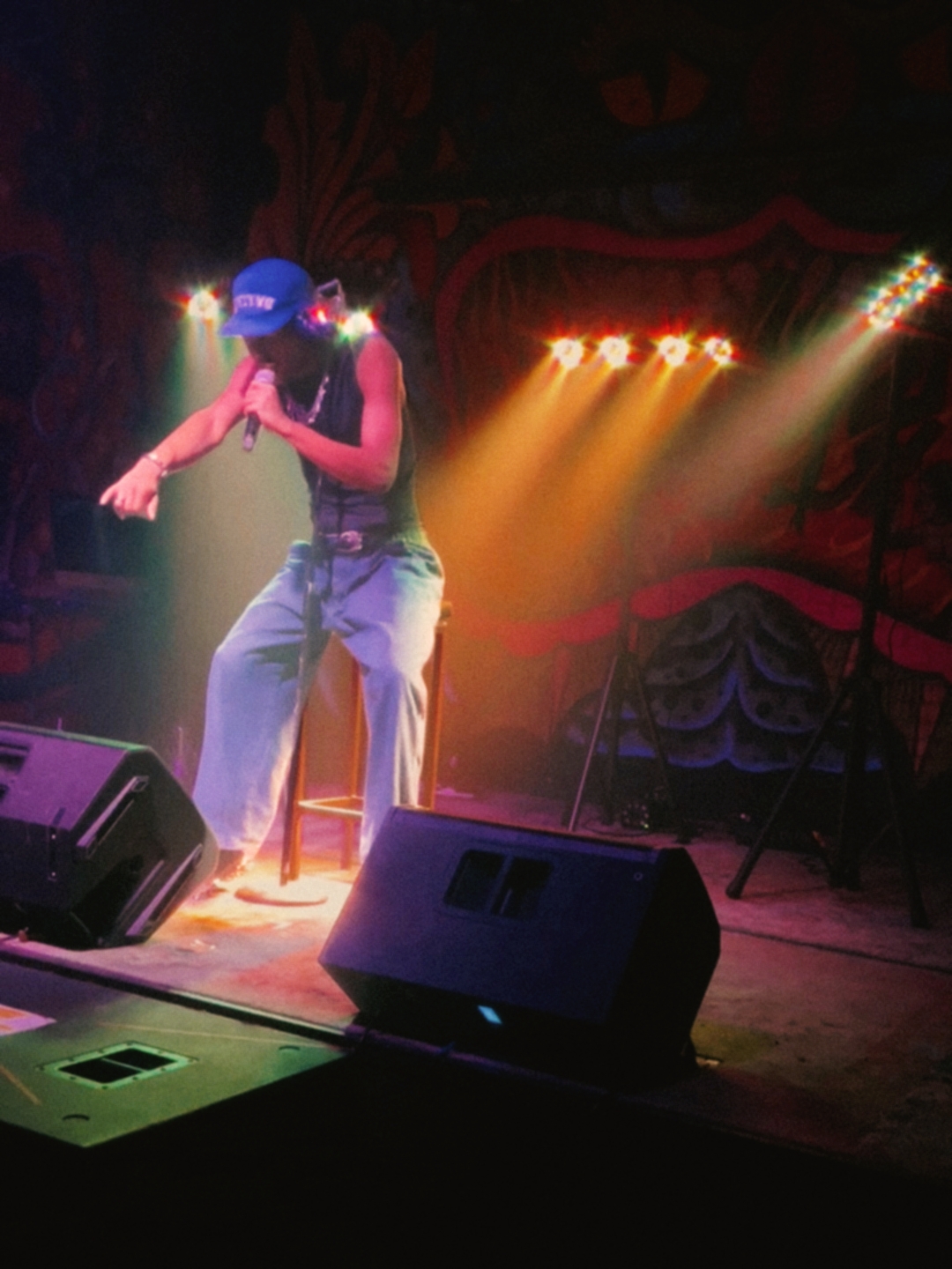 Performer on stage wearing a blue cap, pink top, and light pants, with stage lights and equipment visible.