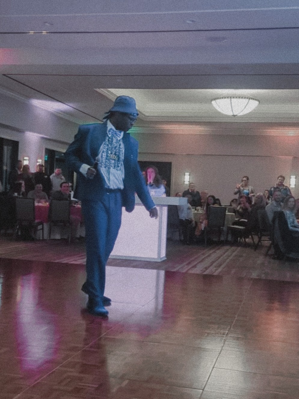 Person in blue suit and hat dancing on a wooden floor in a large event hall with seated guests and a chandelier.