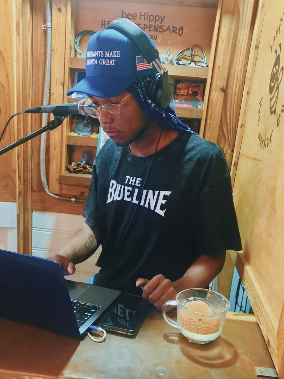 Person wearing a blue cap and headphones, working on a laptop in a wooden room, with a glass of drink nearby.