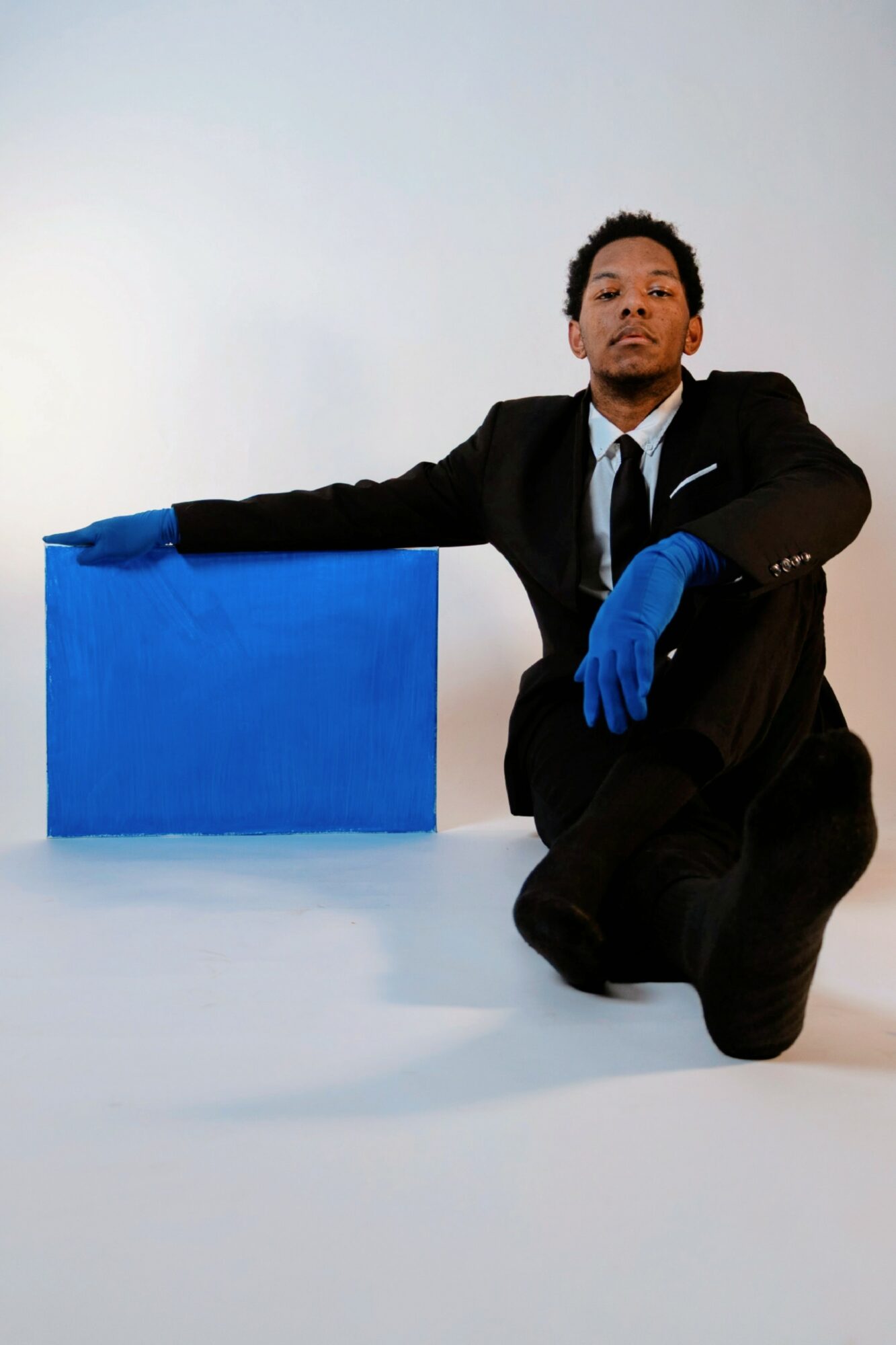 Person in a suit with blue gloves kneeling next to a blue block, against a plain background.