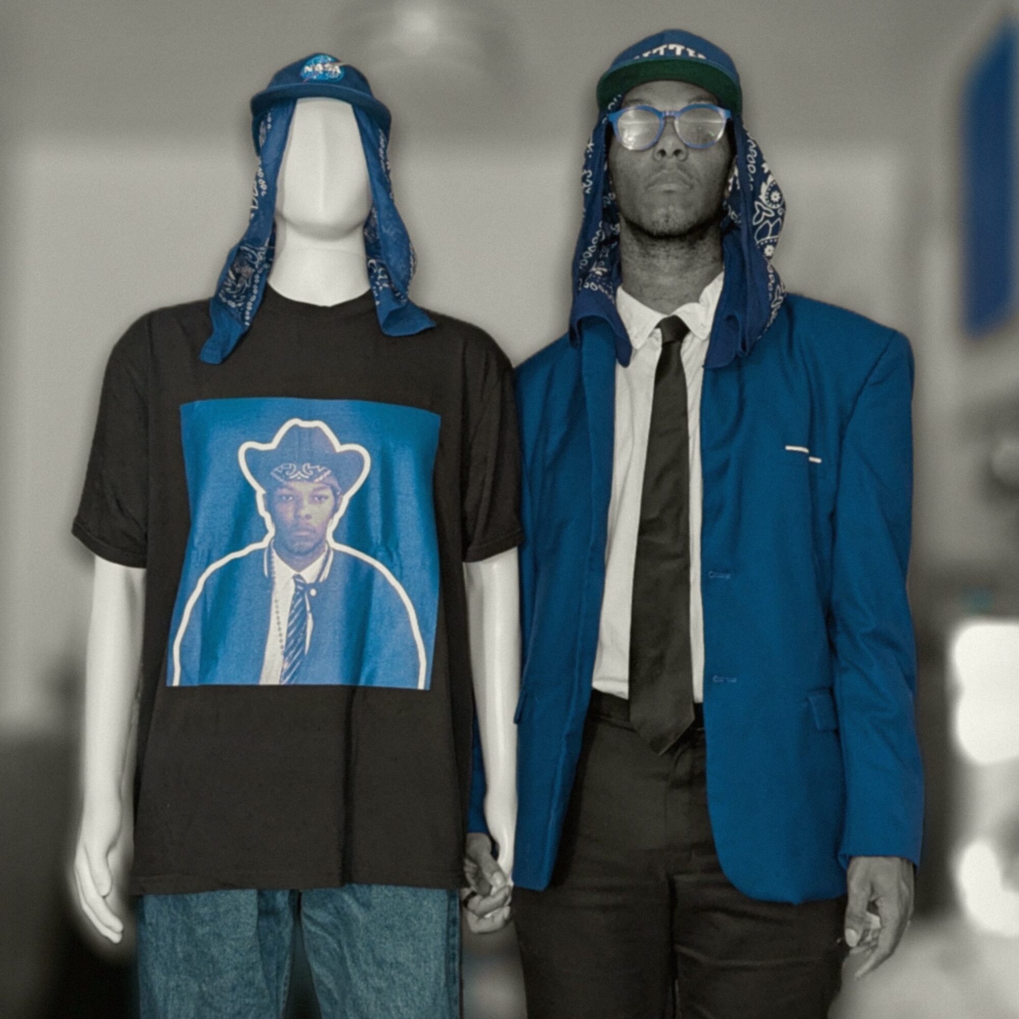 Man and mannequin wearing blue hats, glasses, and casual clothing, standing side by side indoors.