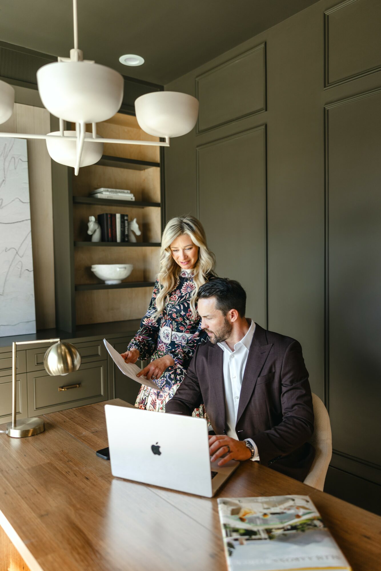 A woman stands behind a seated man working on a laptop in a modern room with dark green walls and wooden furniture.