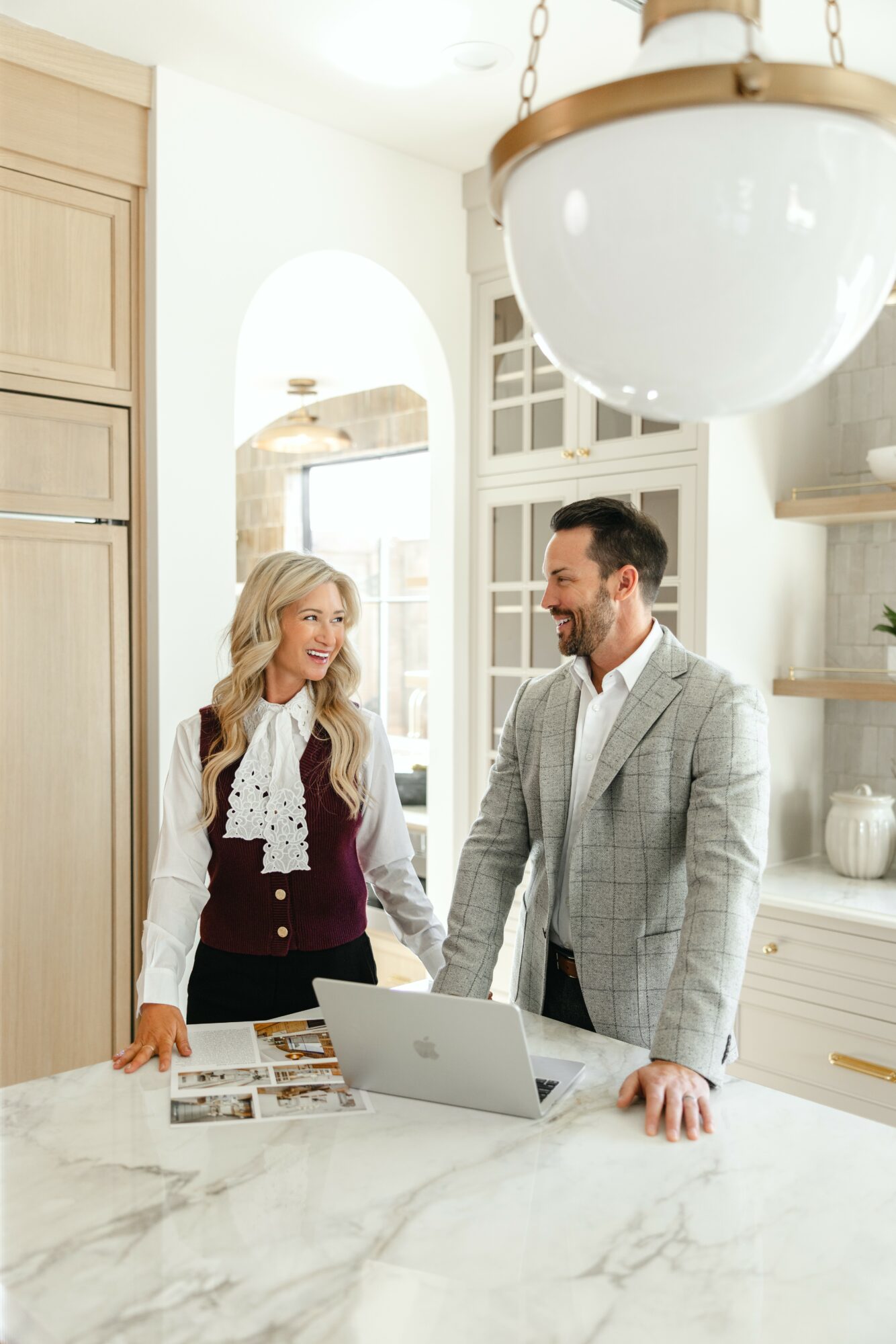Two people, a woman and a man, stand at a kitchen counter with a laptop, smiling at each other.