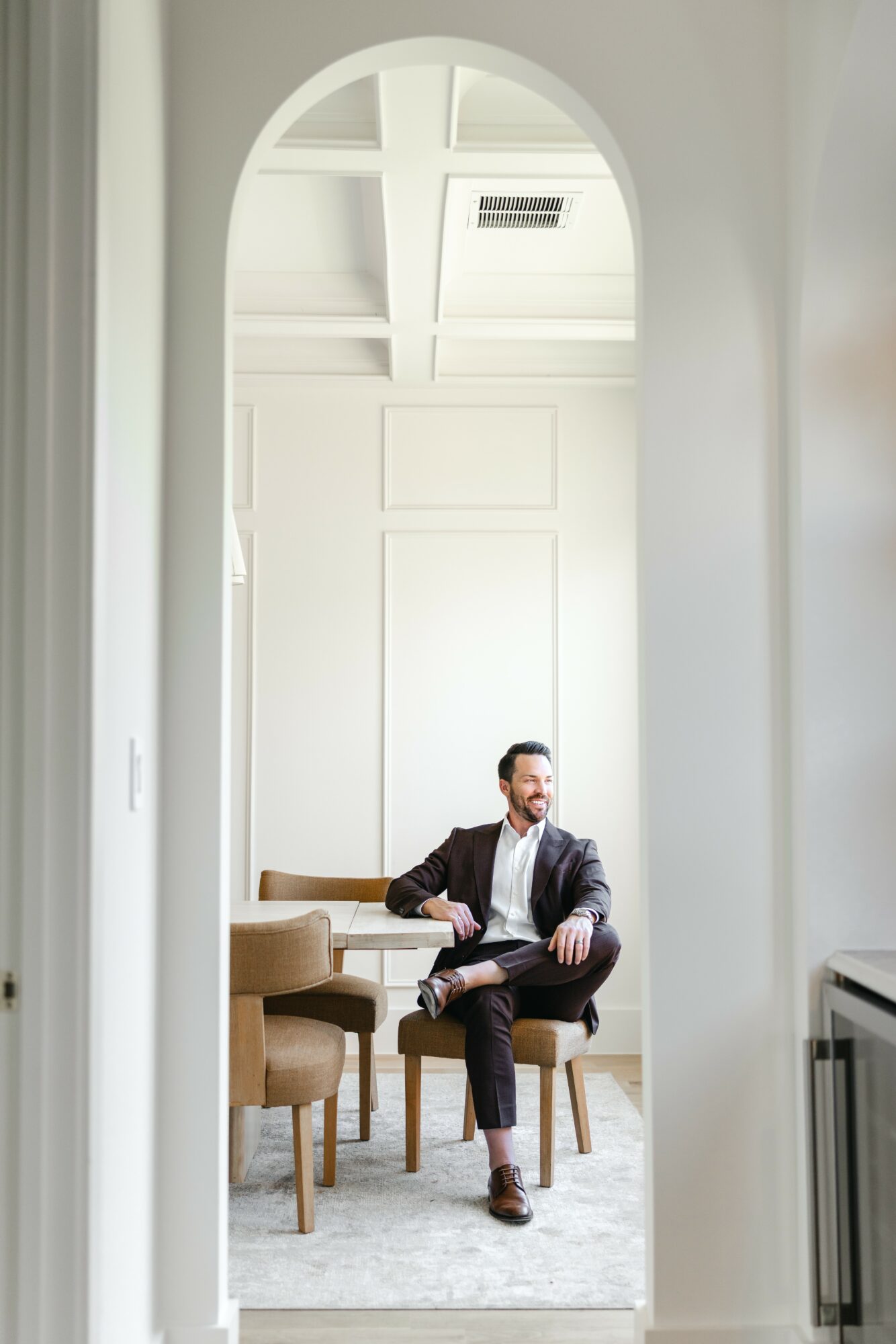Man in suit sitting on a chair in a bright room with high ceiling and white walls.