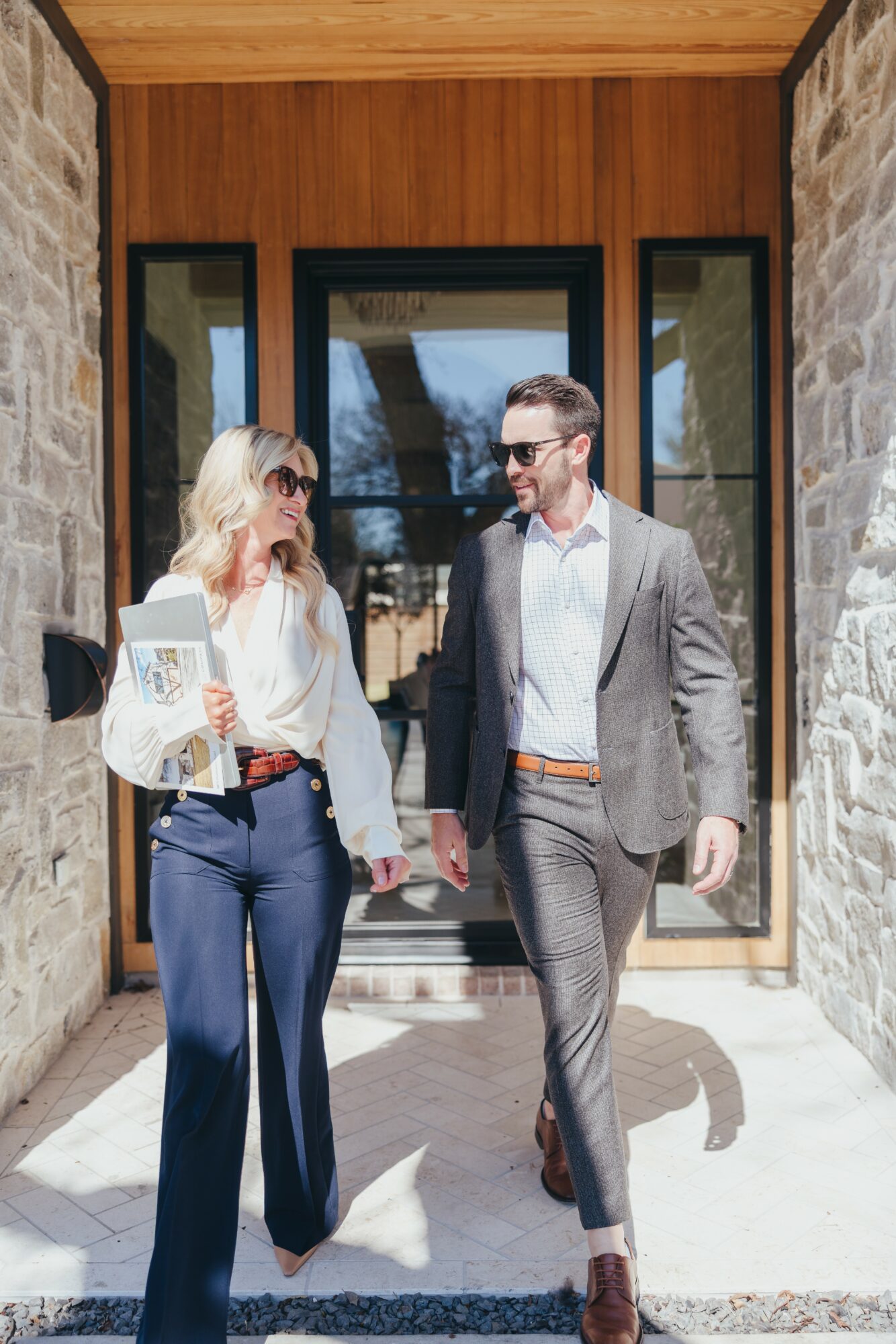 A man and woman walk out of a building with large glass doors, stone walls, and wooden framing, holding hands.