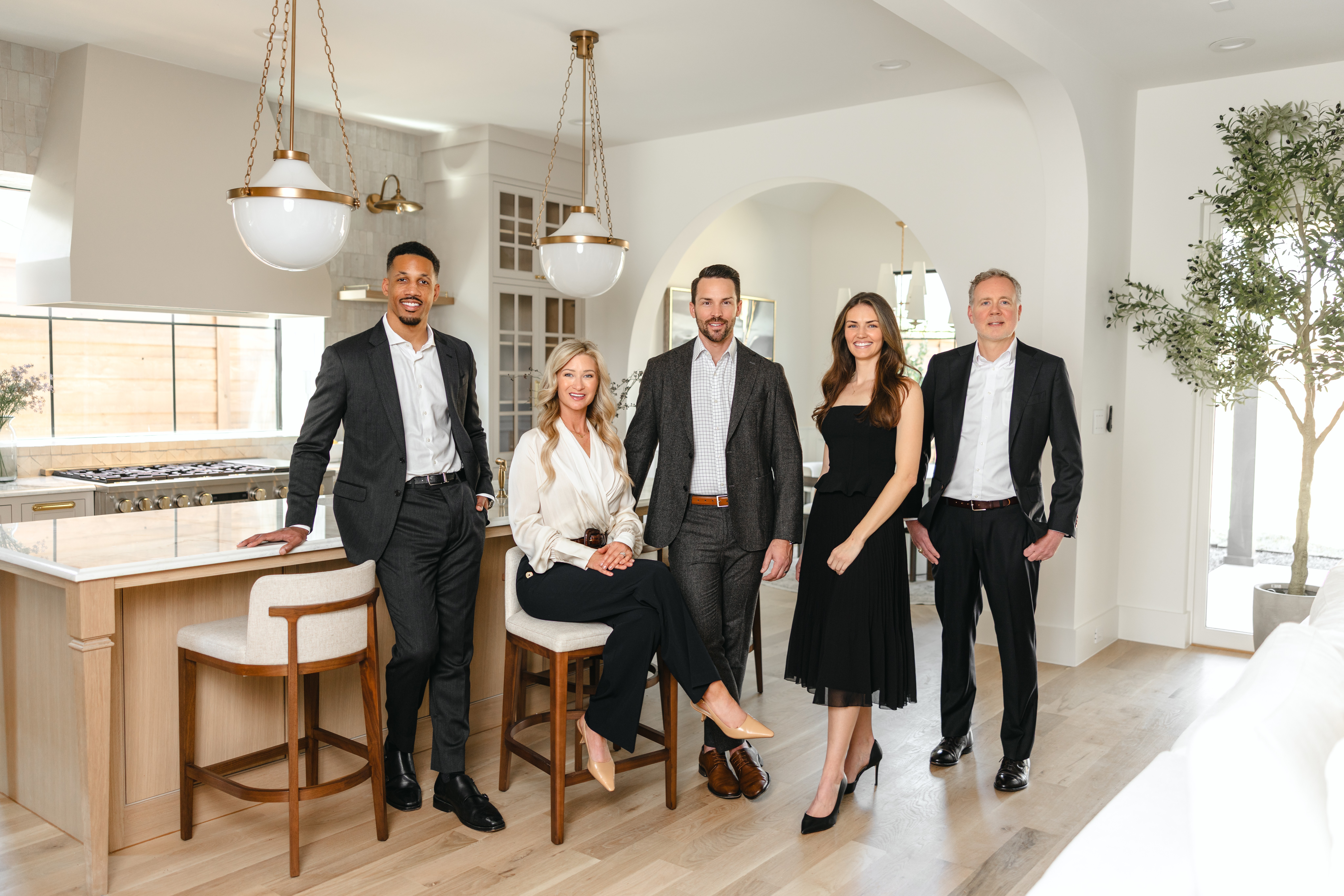 Group of six people in formal attire standing and sitting in a bright, modern kitchen with white walls and wooden floors.