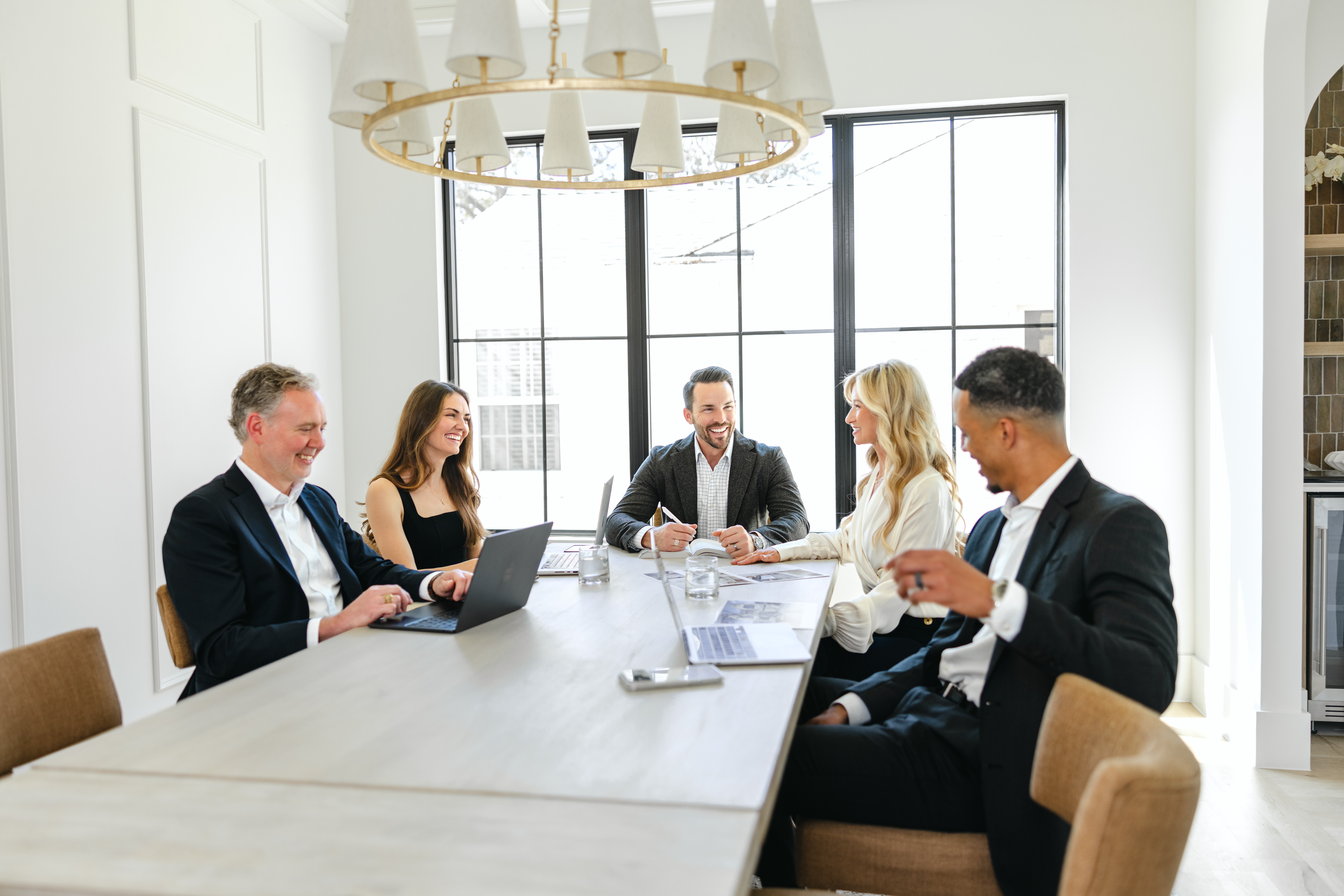 Five people sit around a white conference table in a bright room with large windows, working on laptops and papers.