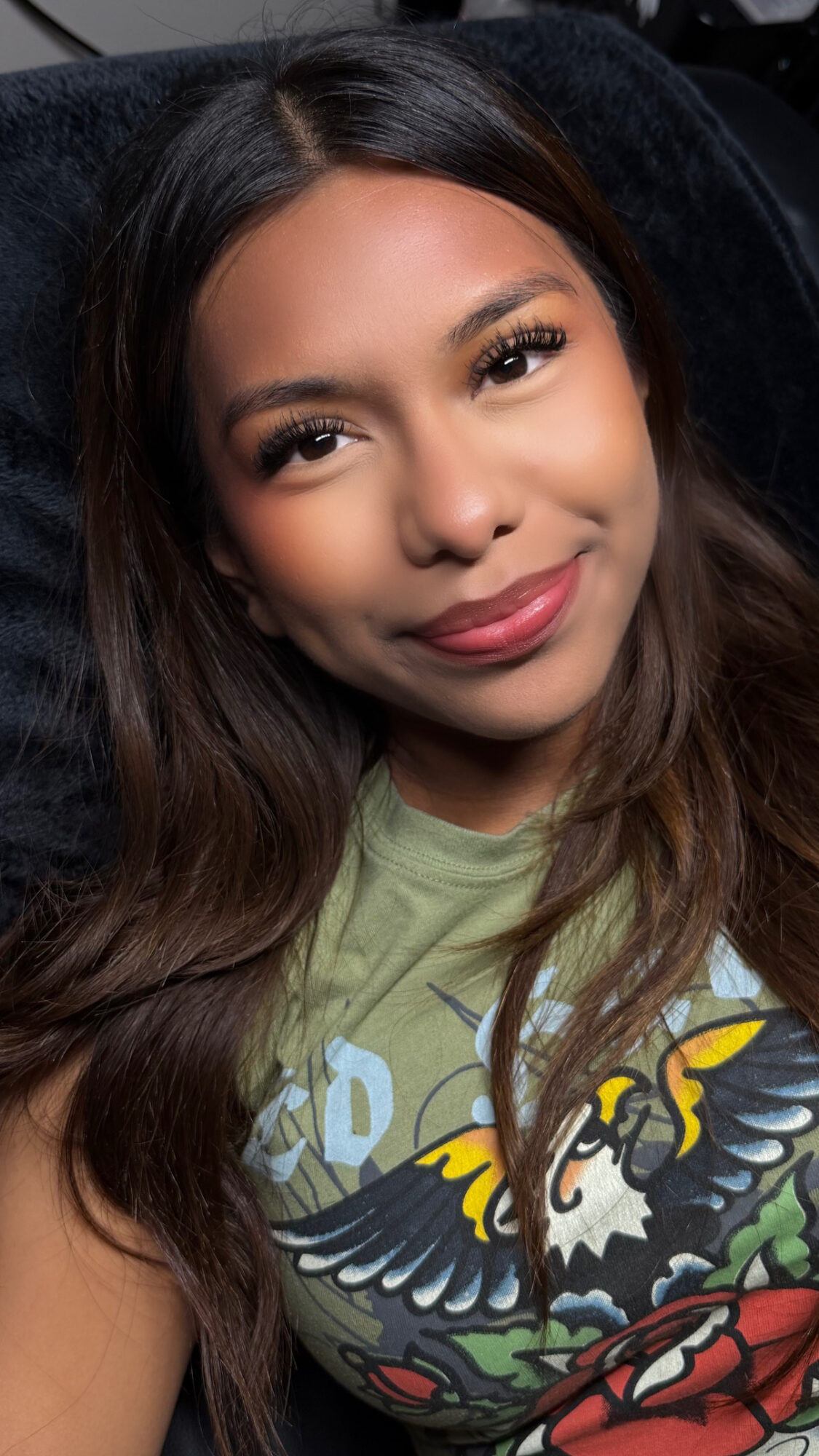 Close-up of a smiling woman with long dark hair, wearing a green shirt with a colorful eagle design.