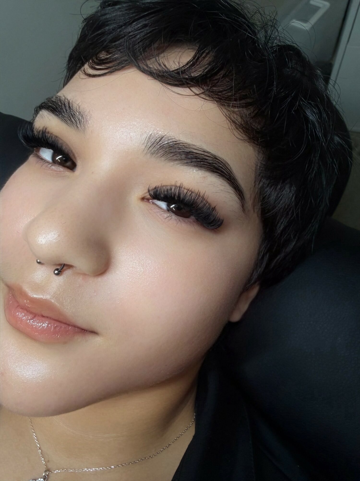 Close-up of a person with short dark hair, makeup, and a septum piercing, smiling slightly.