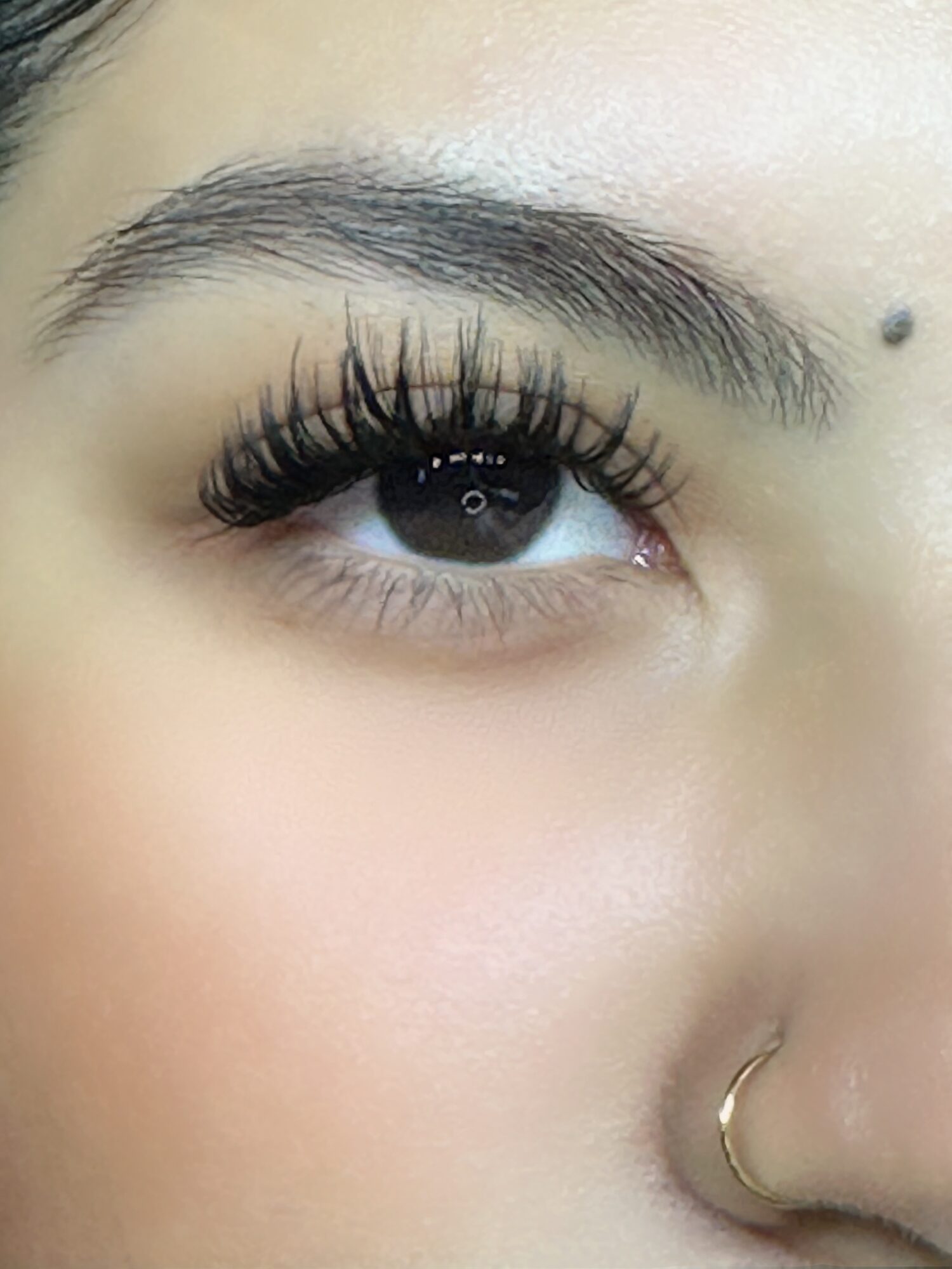 Close-up of a person's eye with long eyelashes, eyebrow, and a nose ring, showing smooth skin and a small mole.