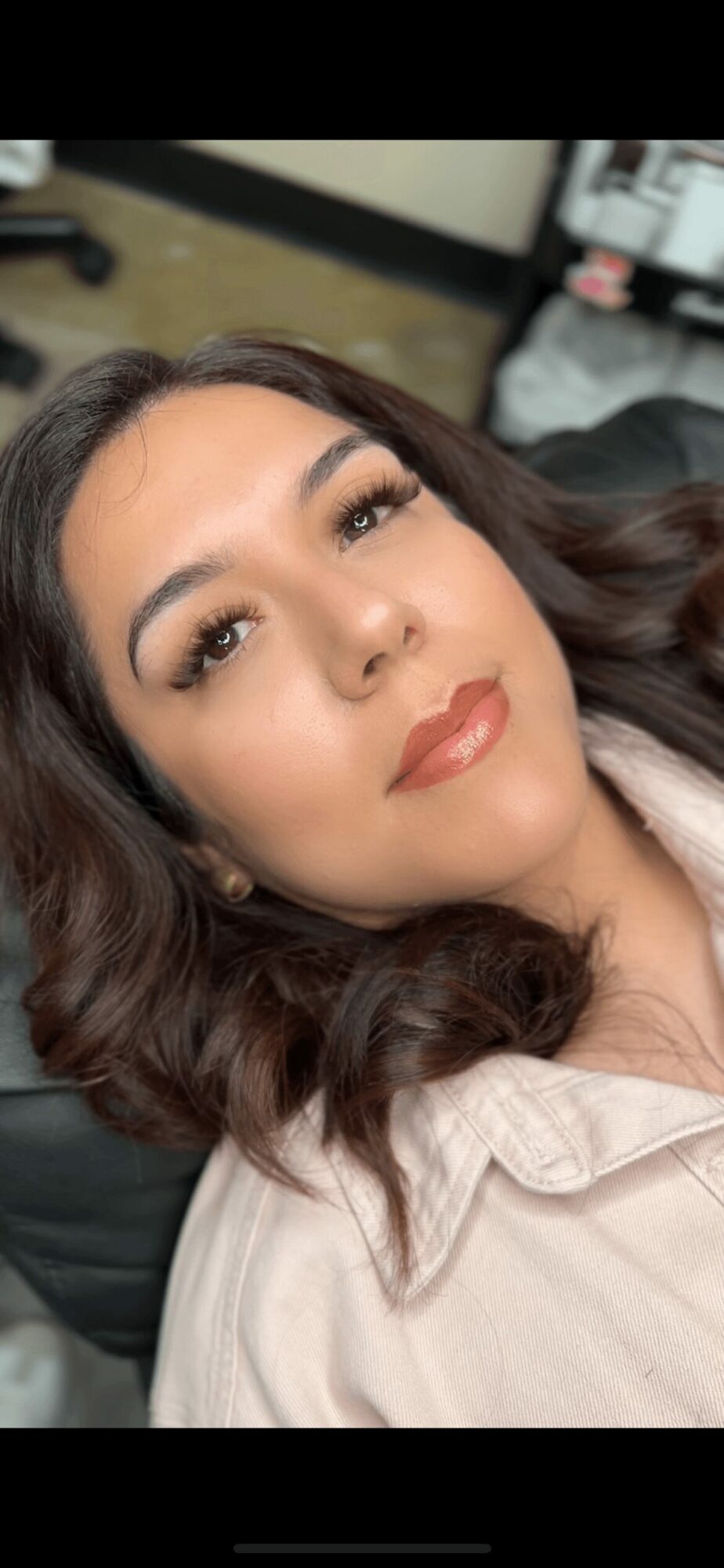 Close-up of a woman with wavy dark hair, makeup, and a light-colored top, lying down indoors.