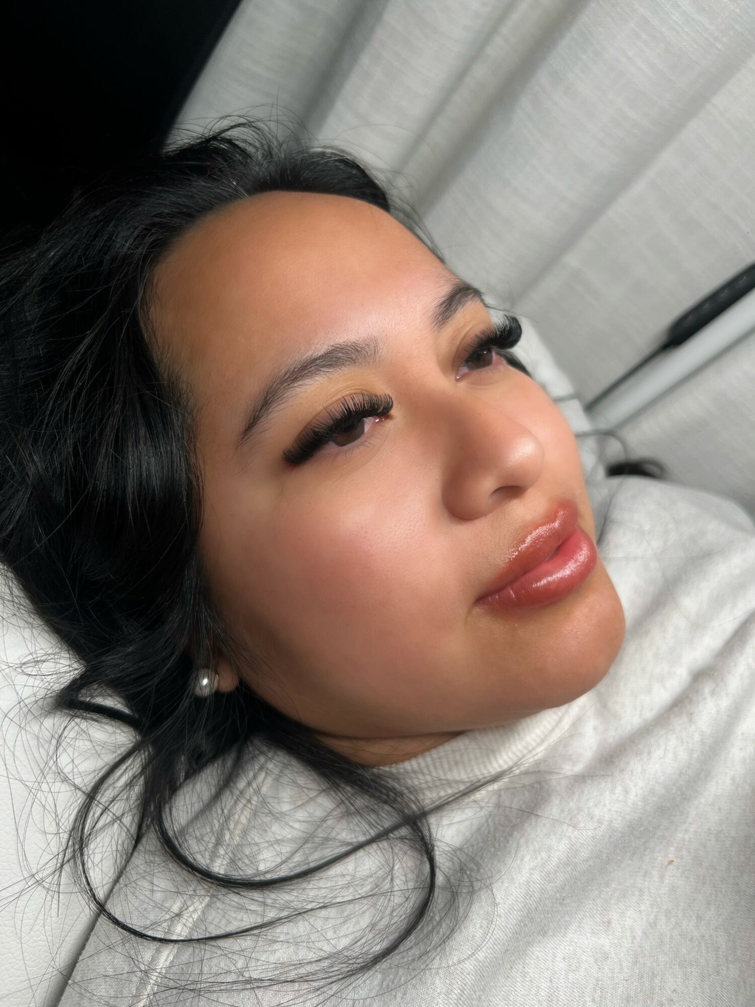 Close-up of a woman lying down with dark hair, makeup, and pearl earrings, looking slightly upward.