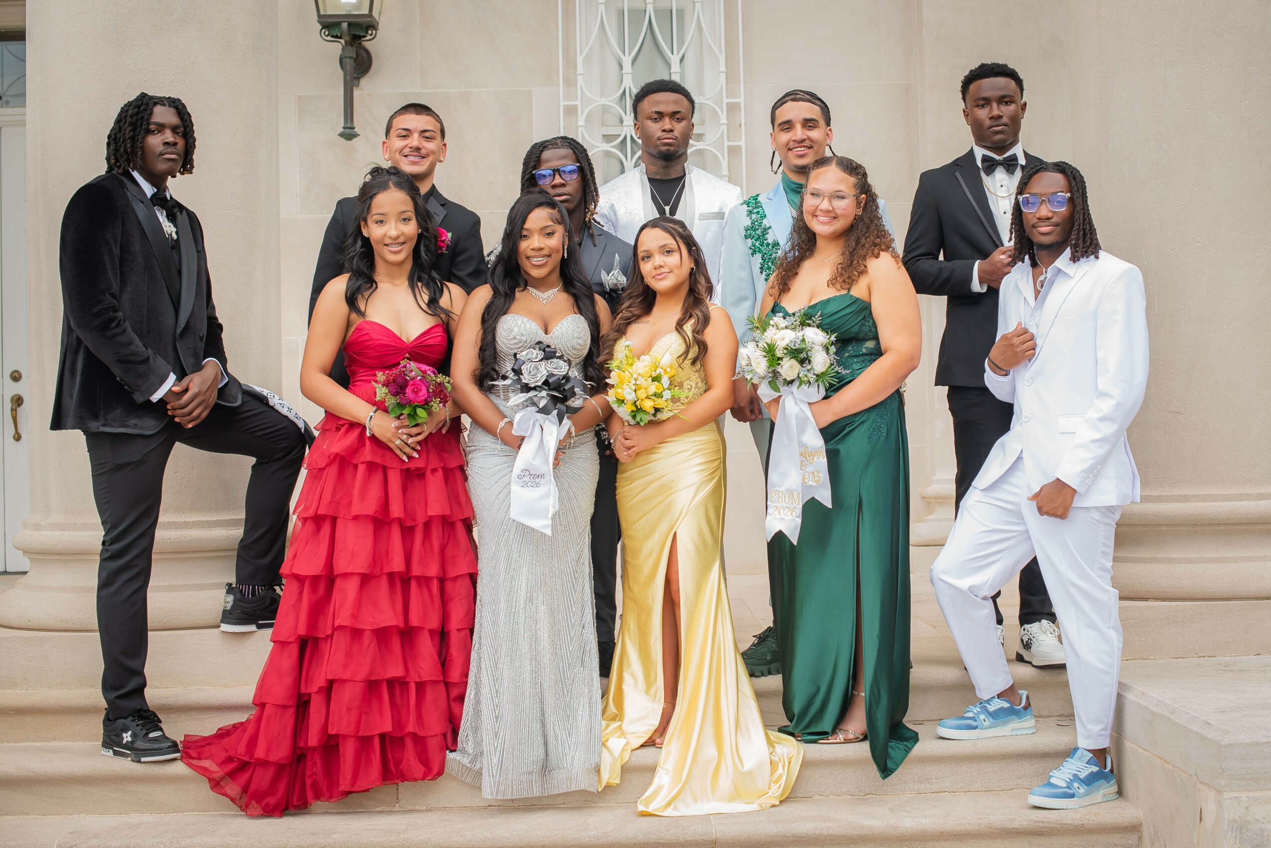 Group of ten people dressed in formal attire standing on steps outside a building, holding flowers, smiling.