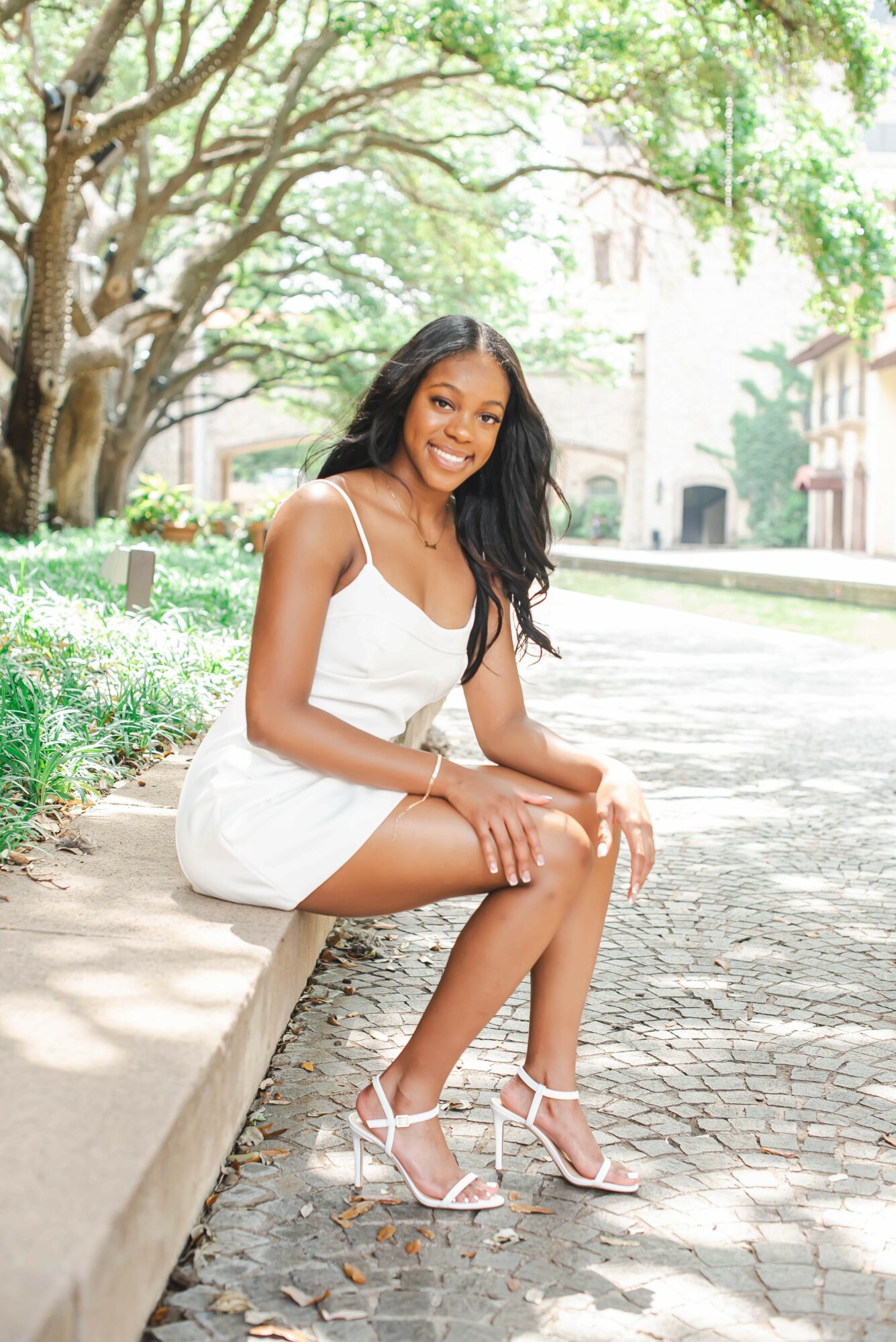 Young woman in white dress and high heels sitting on a park bench, smiling, with trees and buildings in background.