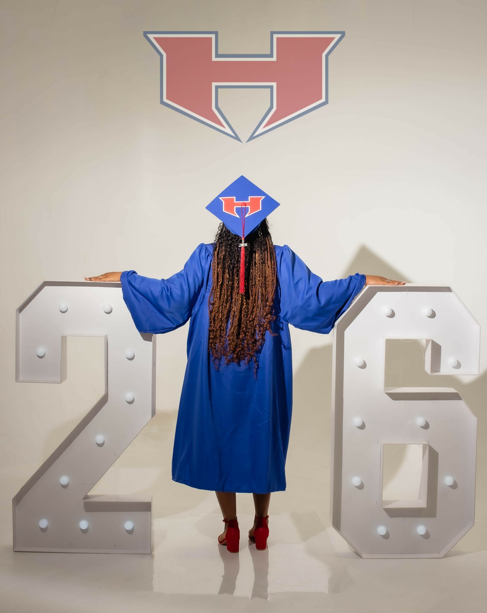 Person in a blue graduation gown and cap standing between large white numbers 2 and 6, with a logo above on the wall.