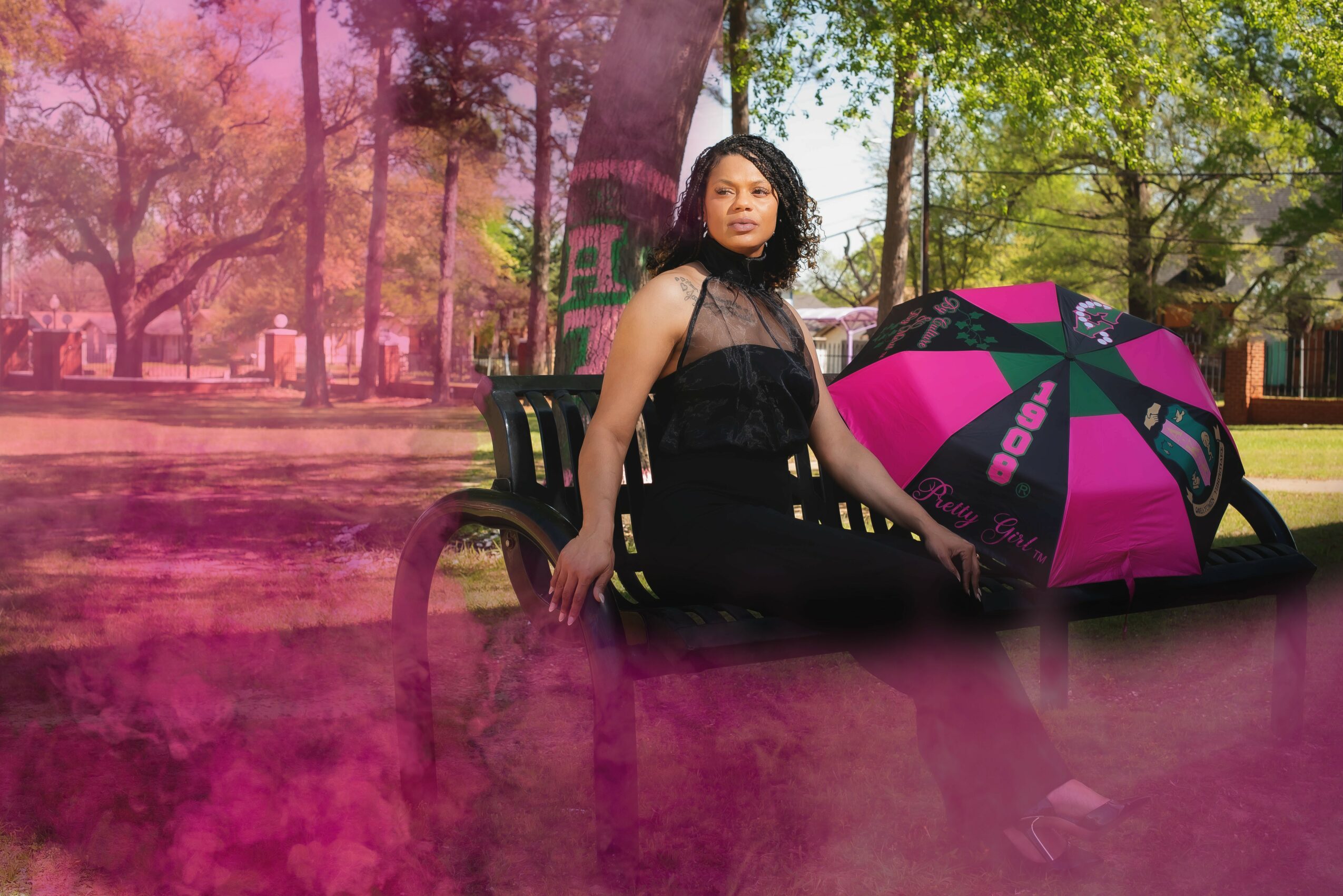 Woman sitting on a park bench with a pink and black umbrella, trees and a pathway in the background, pink smoke in foreground.