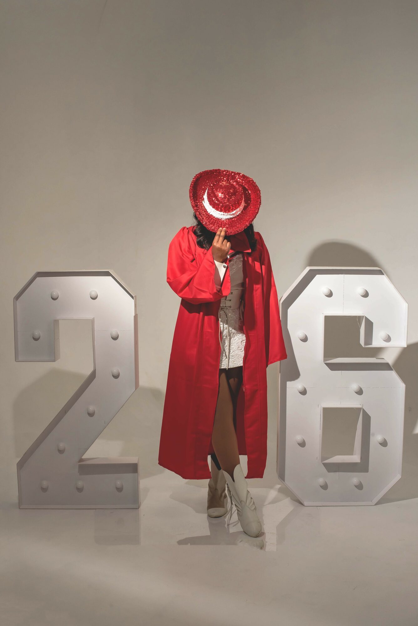 Person in red coat and hat taking a selfie, standing between large white numbers 2 and 6, against a plain background.