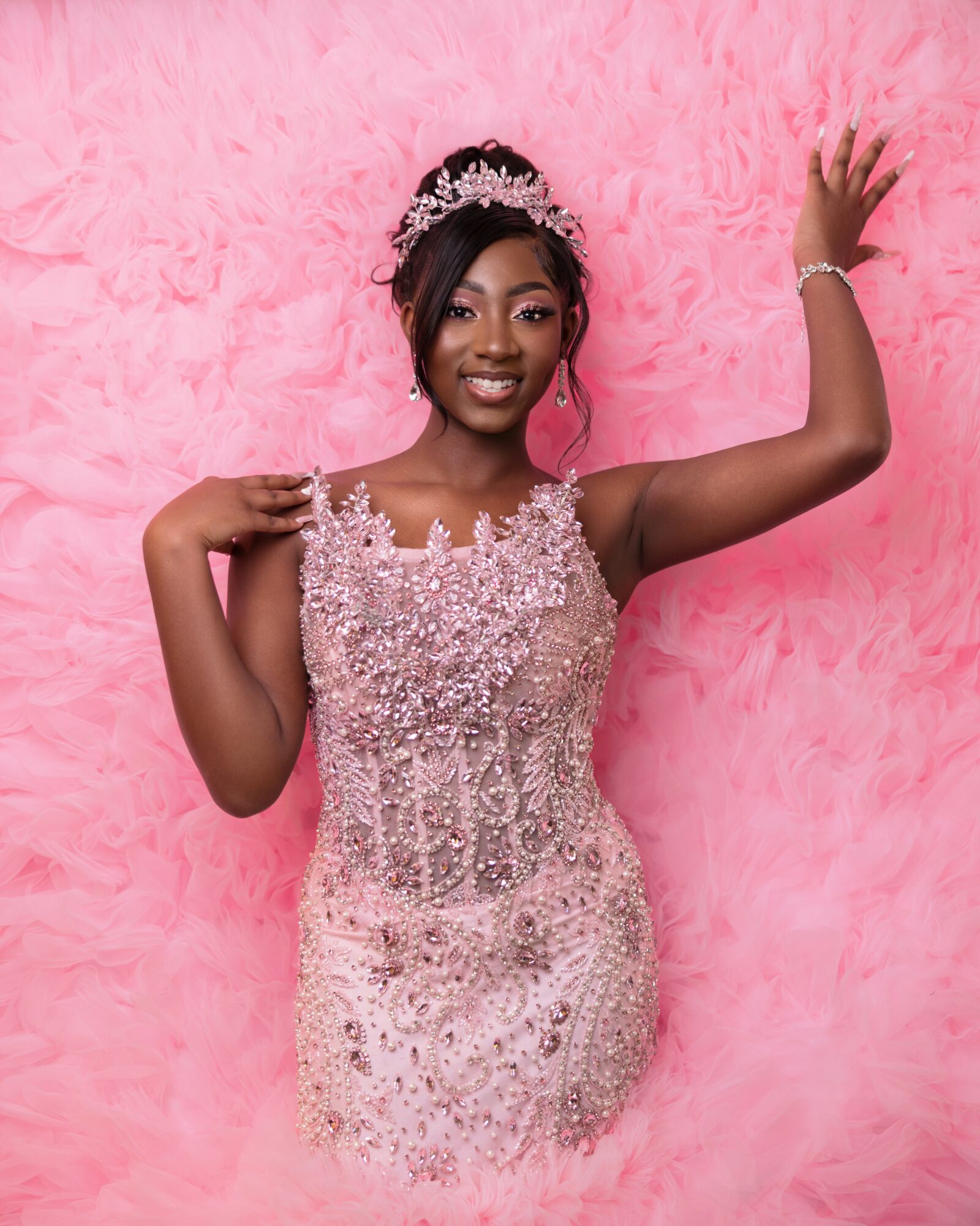 Young woman in a sparkling dress and tiara, smiling, standing against pink fluffy background, waving hand, jewelry visible.