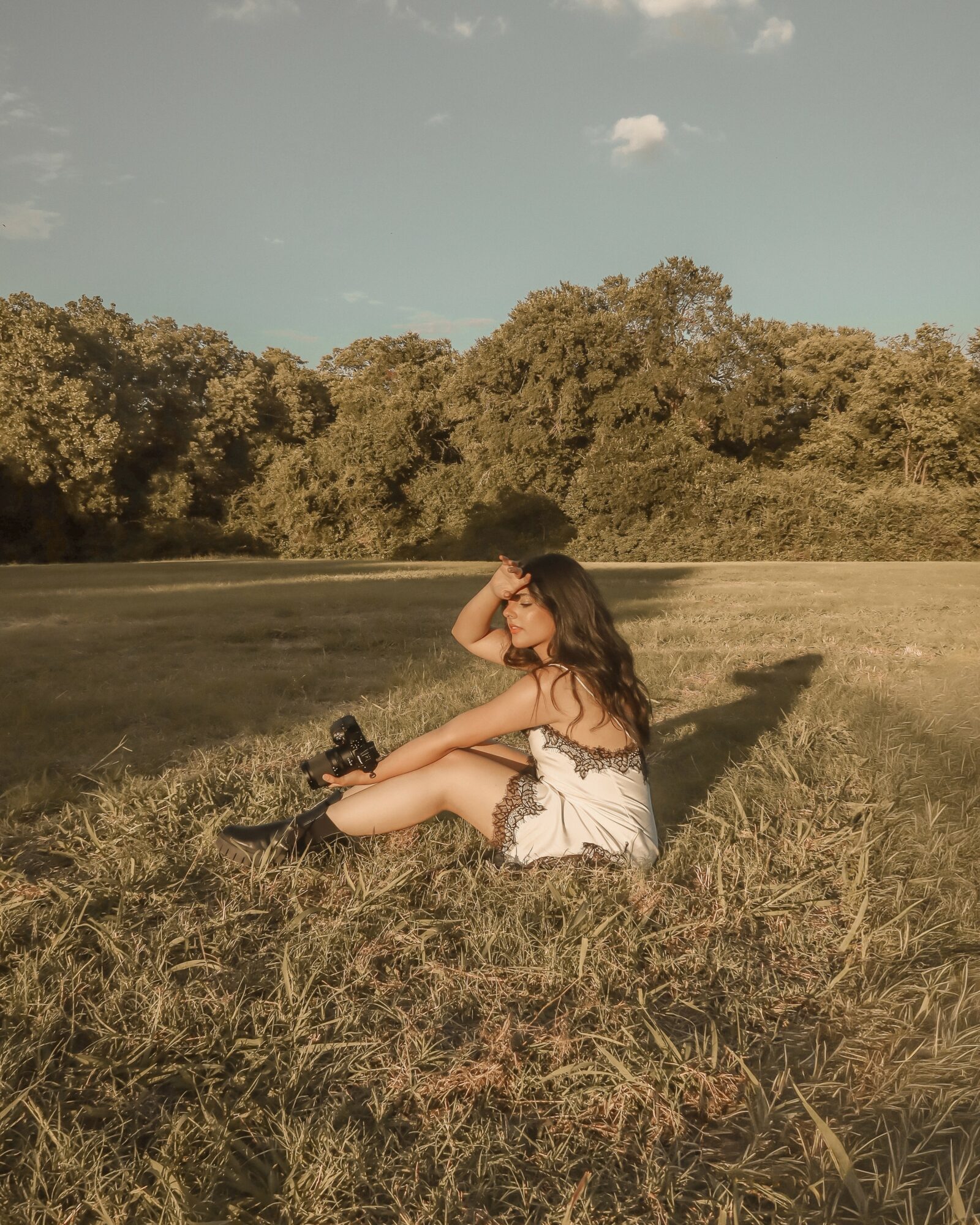 Woman sitting on grass in a field with trees in the background, sunlight casting shadows, wearing a white dress, holding a camera.