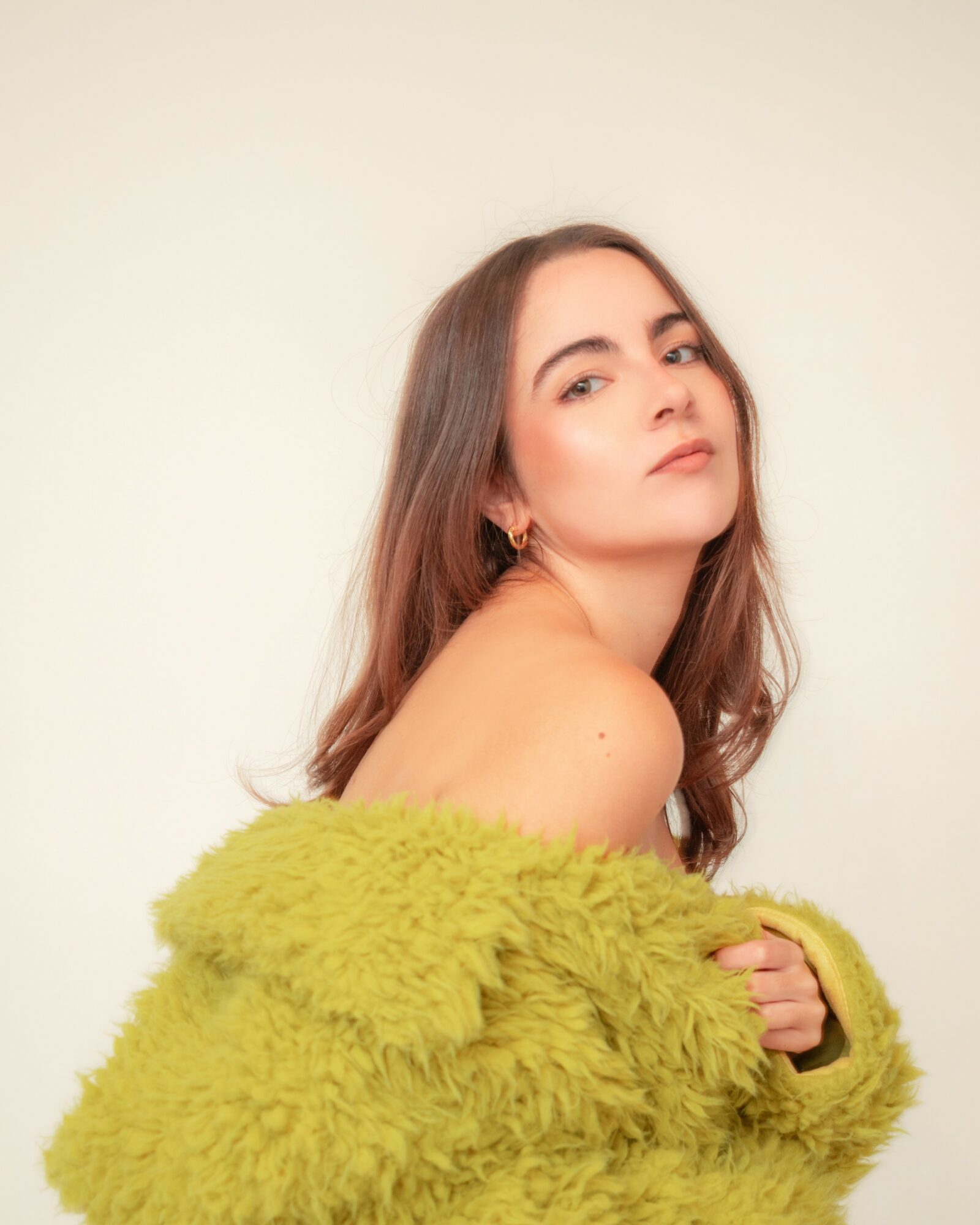 Young woman with long brown hair looking over shoulder, wearing a green fuzzy coat, against a plain light background.