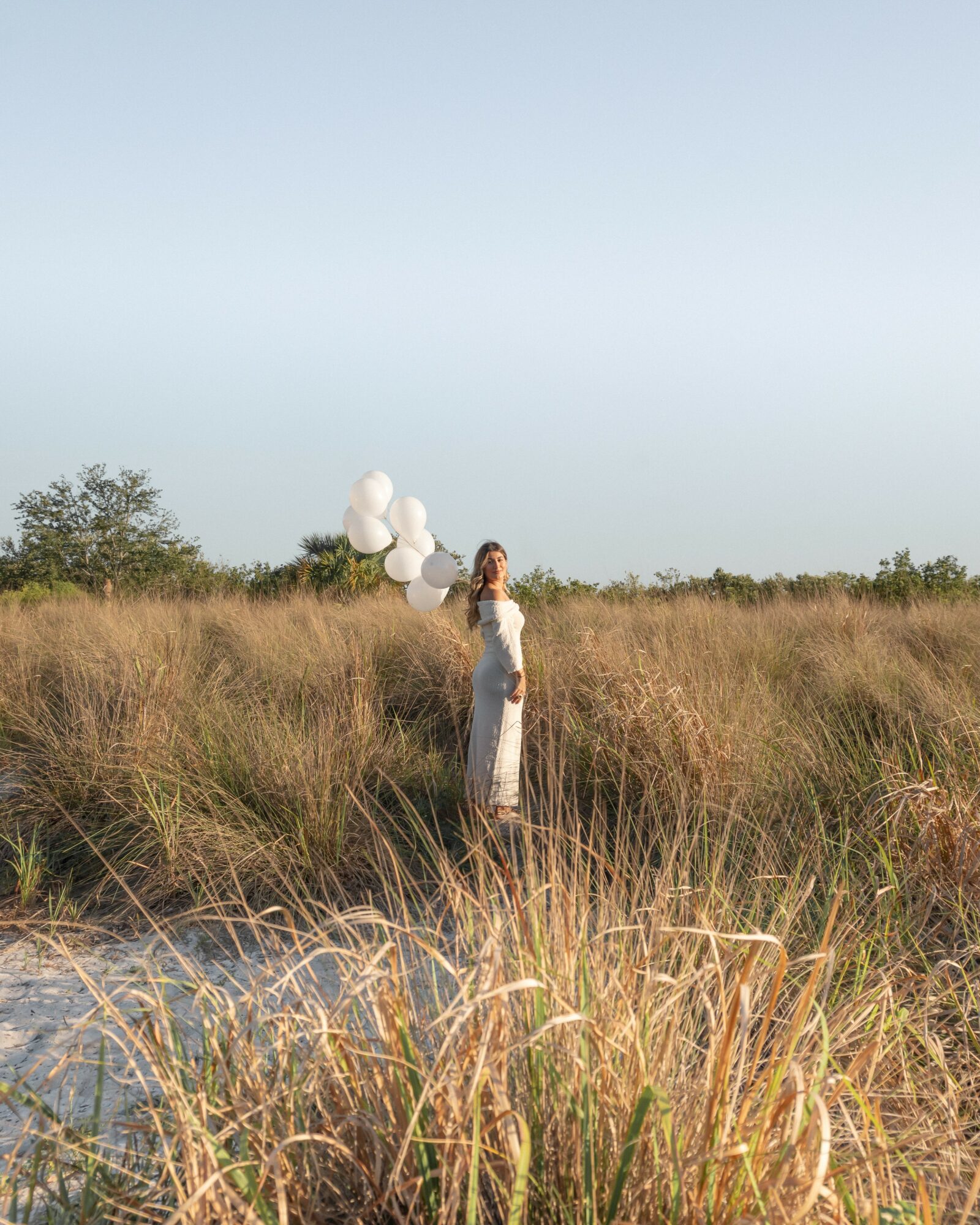 Person standing in a grassy field holding white balloons, with a clear sky and some trees in the background.