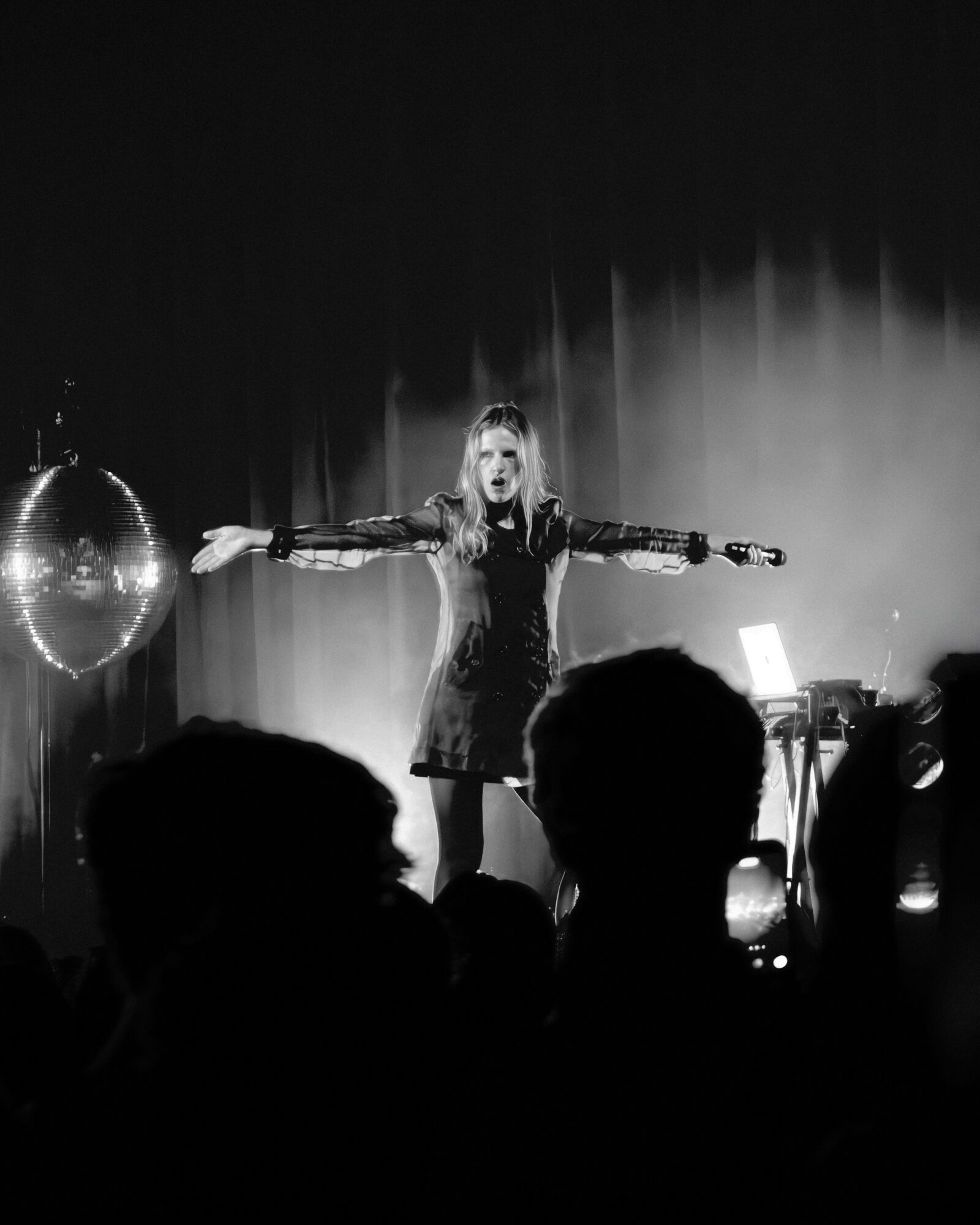Woman on stage with arms outstretched, surrounded by audience silhouettes, in a dark performance setting with a disco ball.