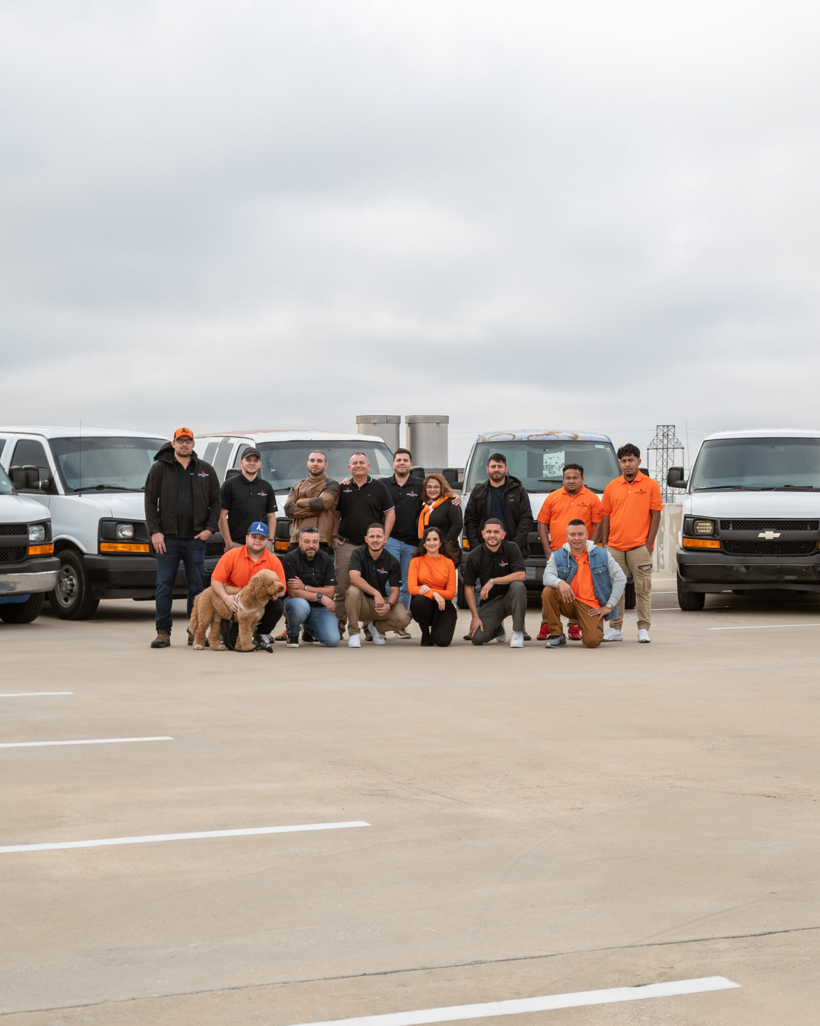 Group of people and dogs in front of vans on a parking lot under cloudy sky.