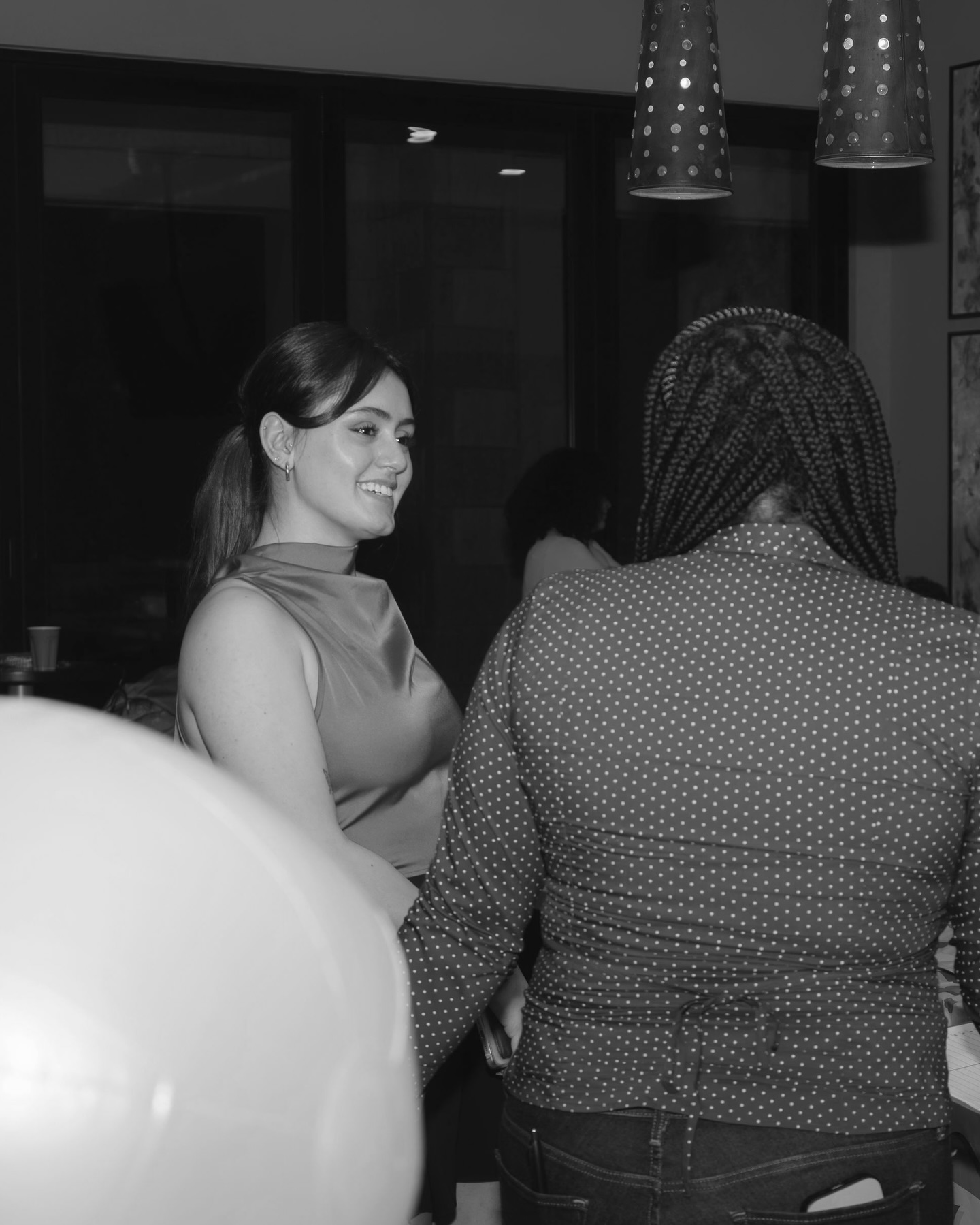 Two women talking indoors, one smiling, with hanging lights above. Black and white photo, background shows windows and decor.