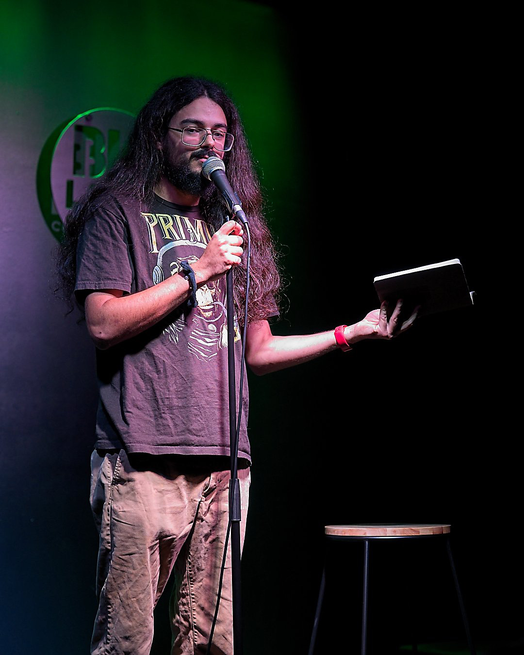 Person with long hair and glasses holding a microphone and a tablet on stage with a green background.